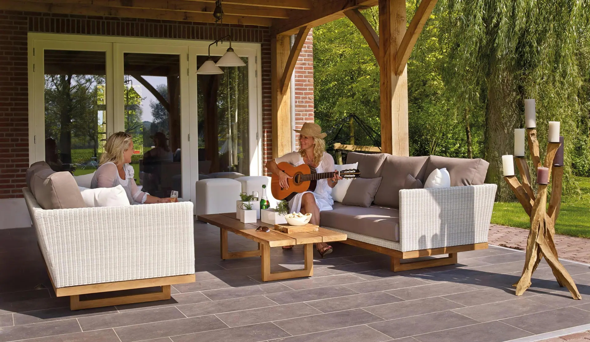 A cozy outdoor setup highlighting patio decor ideas with wicker sofas with wooden elements, soft neutral cushions, and a matching low wooden coffee table. Two women relax under a covered patio - one playing guitar, the other enjoying a drink, surrounded by greenery and warm natural light, creating an inviting and tranquil ambiance.