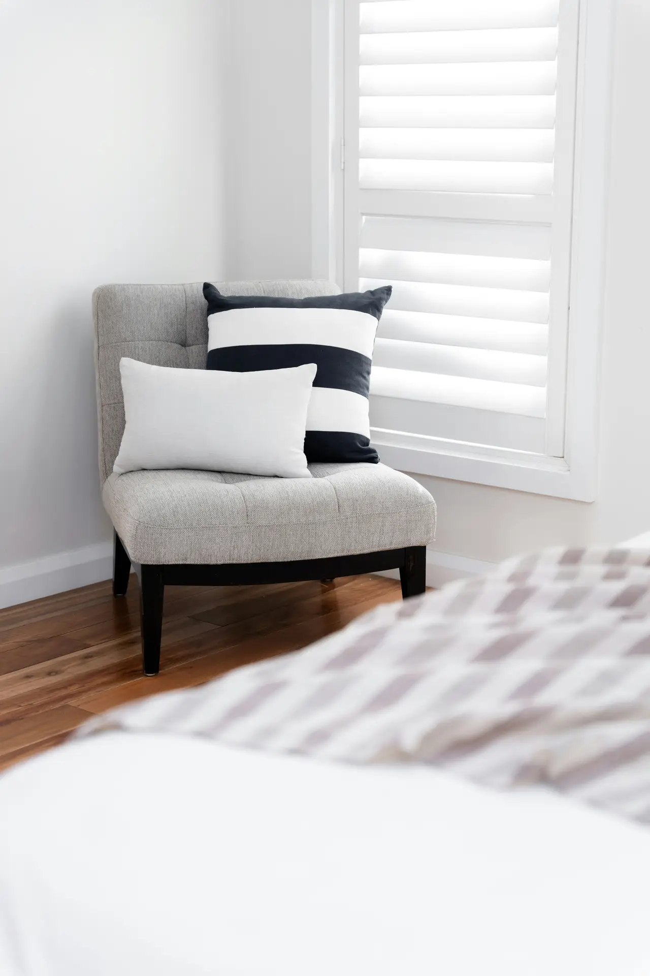 A bright bedroom corner featuring a cozy corner chair upholstered in soft gray fabric, styled with a striped cushion and a simple white pillow. Sunlight filters through modern shutters, adding to the calm mood and offering bedroom ideas for creating a comfortable little nook within a minimalist space.
