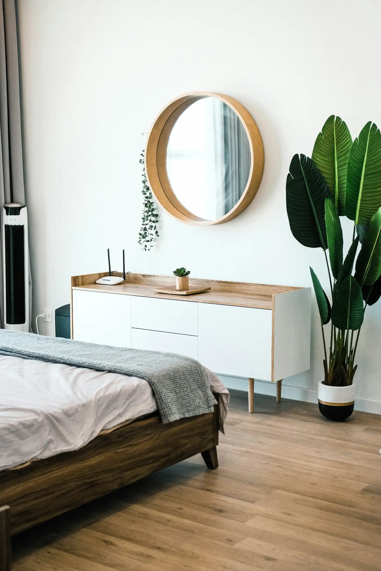 A bright bedroom with warm wood floors, a simple bed frame, and a sleek sideboard topped with decluttered surfaces, complemented by a round mirror and a tall indoor plant, an inspiring setup for bedroom ideas that balance calm design with effortless organization.