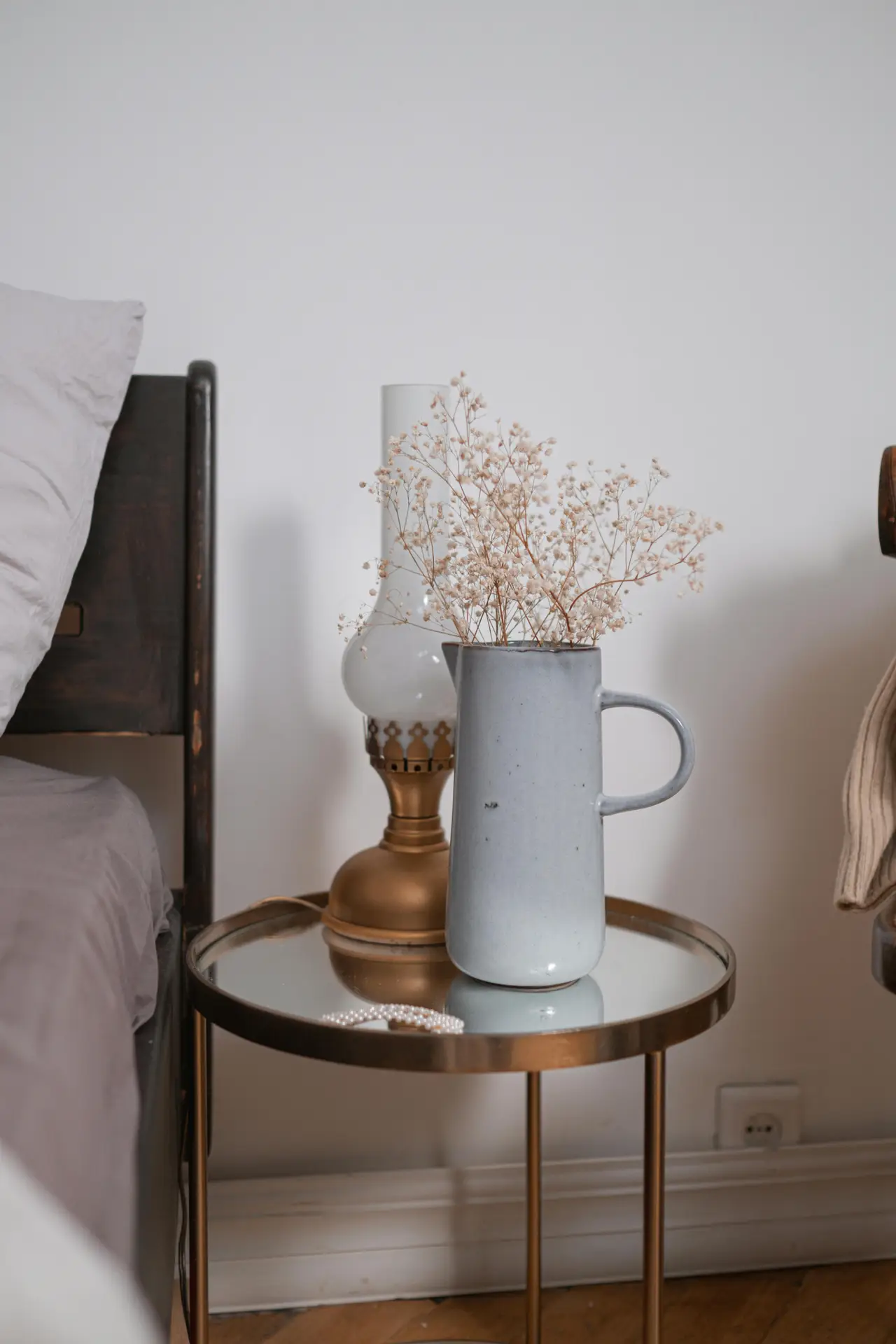 A simple bedside setup featuring a round glass-top table with subtle metal accents, holding a ceramic pitcher with dried flowers and a vintage-style lamp, offering relaxed bedroom ideas that blend soft textures with a hint of shine.
