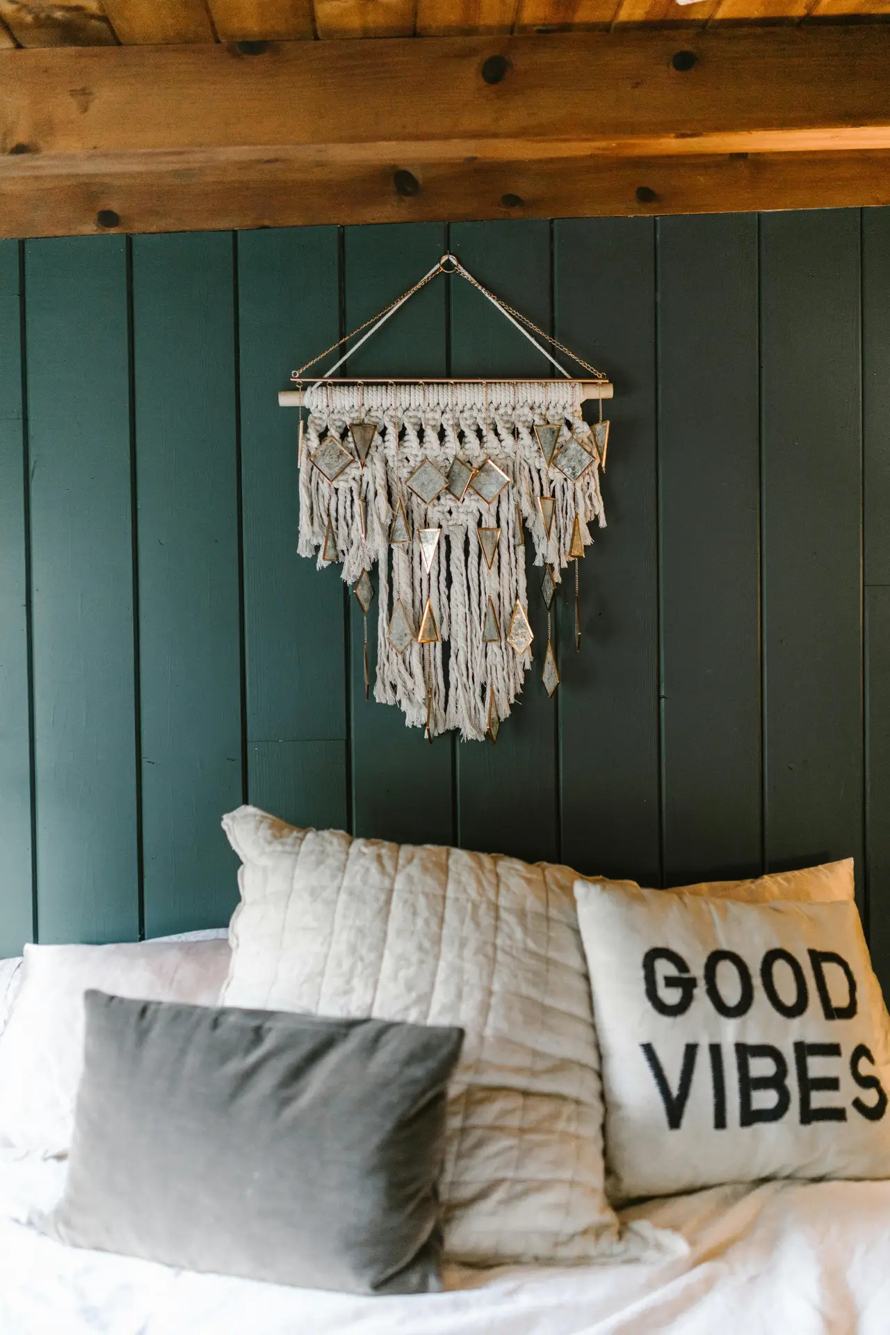 A cozy bedroom setup with soft layered pillows against a dark green paneled wall, accented by a macramé-style hanging that adds minimal wall decor without overwhelming the space. Warm wood beams overhead and relaxed textures give inviting bedroom ideas for creating a calm, rustic-inspired atmosphere.