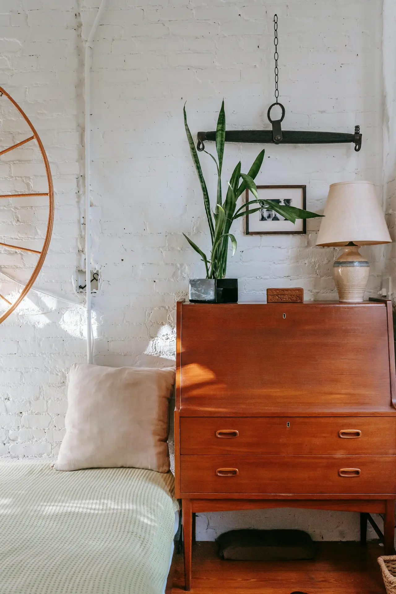 A bright bedroom corner featuring a vintage wooden dresser topped with a lamp and a tall plant, placed against a white brick wall. A simple bed with a soft pillow sits beside it, creating a calm setup that highlights bedroom ideas built around a mix of old and new pieces for a warm, collected feel.
