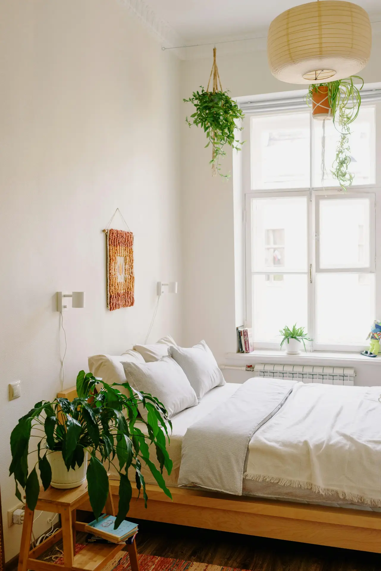 A bright bedroom filled with natural greenery, featuring a simple wooden bed layered with soft neutral bedding and pillows. Hanging plants, potted leaves on a small stool, and a few accents by the window show how bedroom ideas can incorporate fresh, organic touches to create an inviting and calming atmosphere.