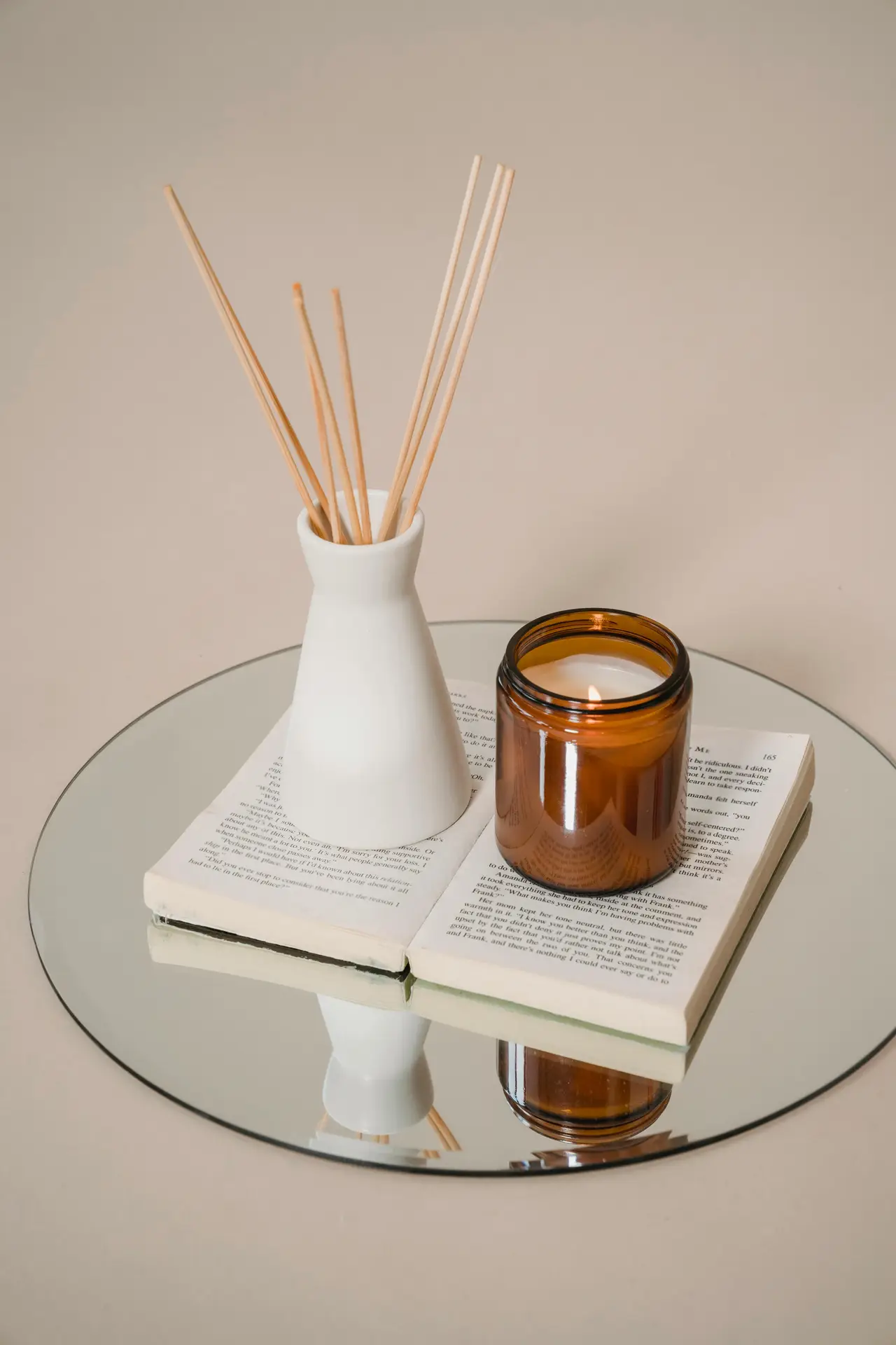A small arrangement of bedroom accessories featuring a lit amber candle and a ceramic diffuser with wooden reeds placed on an open book atop a round mirror, offering simple bedroom ideas that incorporate natural scents for a calm and inviting atmosphere.