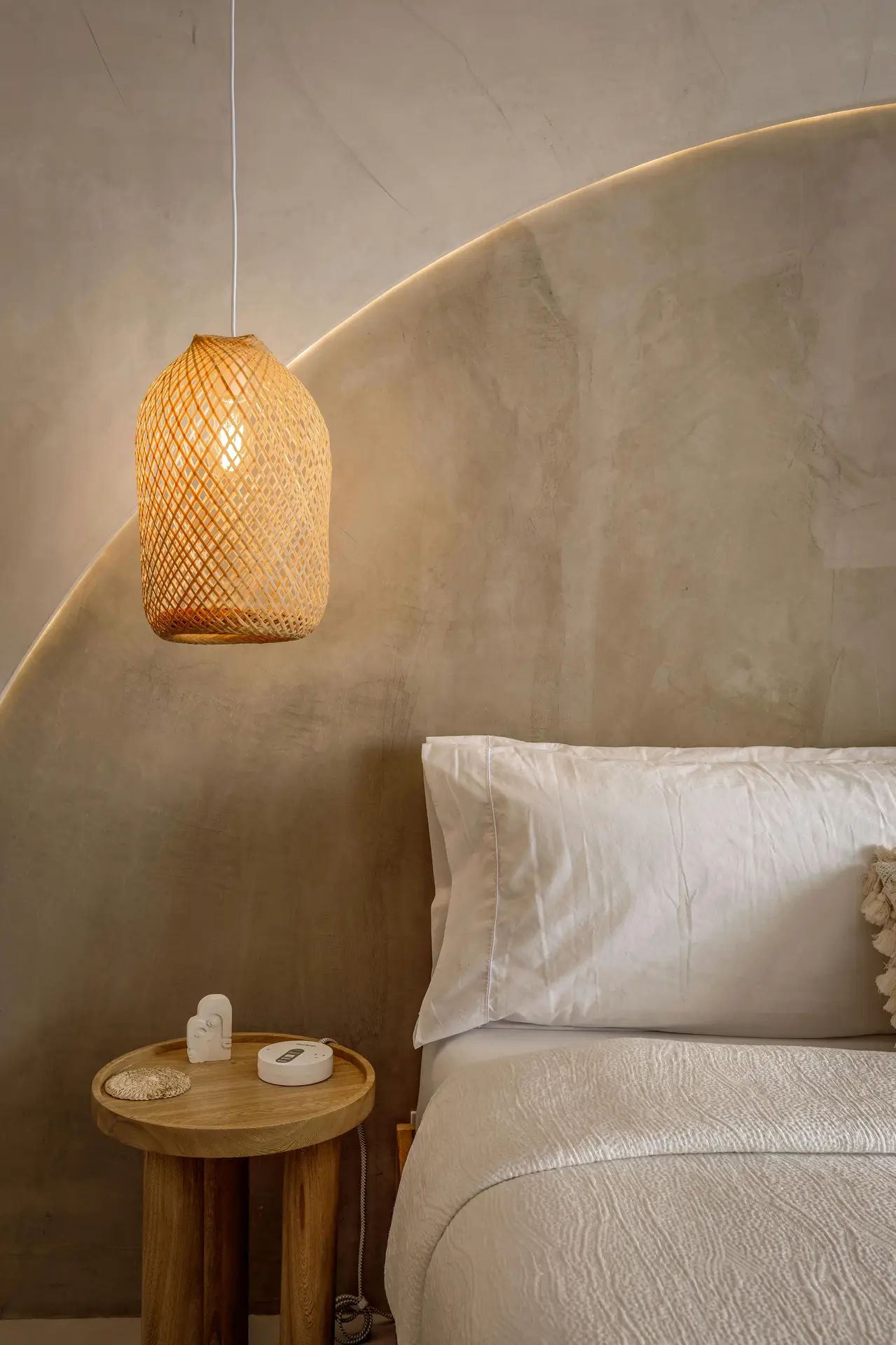 A serene bedroom scene featuring a neutral-toned wall with a curved LED glow, offering bedroom ideas that highlight soft layered lighting. A woven pendant lamp casts a warm ambiance above a simple wooden bedside table, next to a neatly made bed with crisp white bedding for a calm, understated look.