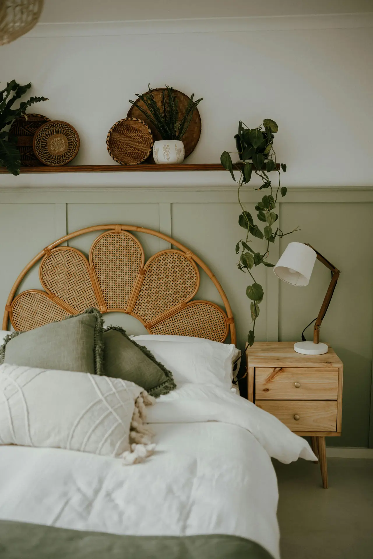 A calm bedroom scene featuring a natural rattan statement headboard shaped like petals, styled with soft green and white bedding for fresh bedroom ideas. A wooden nightstand with a simple lamp and a trailing plant adds warmth, while woven baskets and greenery on the shelf above create an earthy, collected look.
