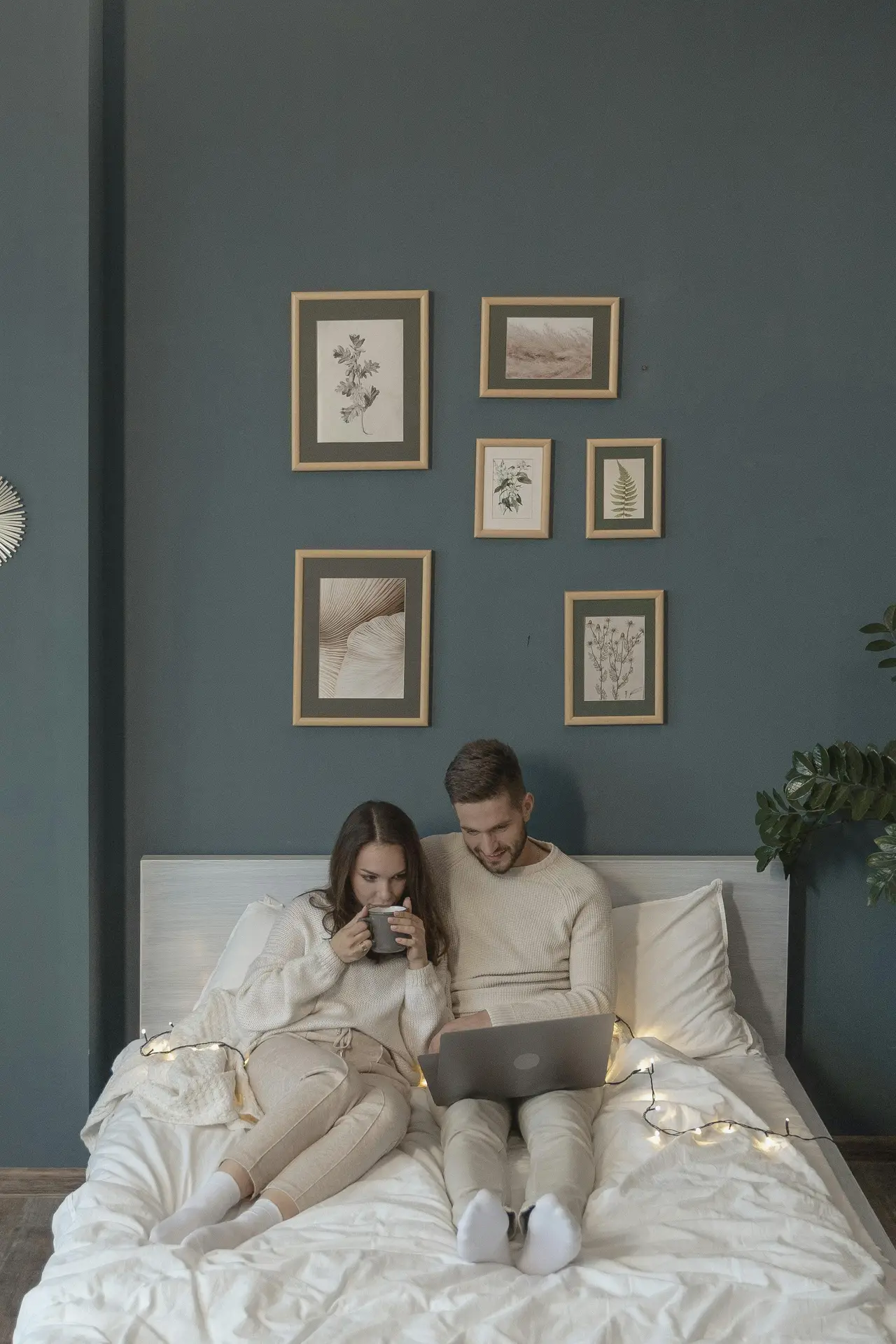 A cozy bedroom setup featuring a couple relaxing on a white bed, surrounded by soft bedding and warm string lights. The dark blue wall behind them is decorated with tonal artwork in simple wooden frames, adding calm visual interest and inspiring bedroom ideas for creating a serene, layered space.