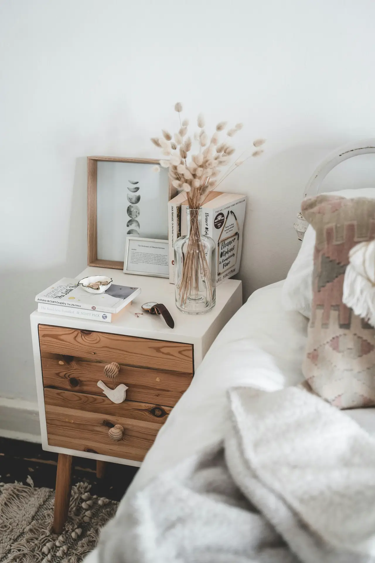 A soft and inviting bedroom setup featuring a compact wooden nightstand topped with books, dried florals in a glass vase, and small decorative pieces, offering bedroom ideas that highlight the charm of wooden nightstands. The edge of a cozy bed with textured pillows and a warm throw blanket adds to the relaxed, lived-in feel.
