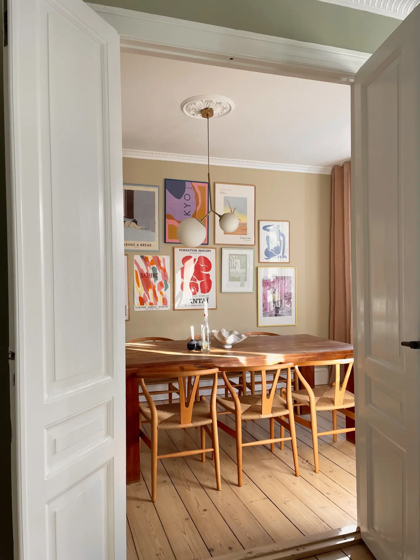 A warm dining space viewed through open white doors, featuring a walnut table with woven chairs and an accent wall in beige decorated with colorful framed artwork, creating a look that blends cozy living rooms with a welcoming, art-filled atmosphere.