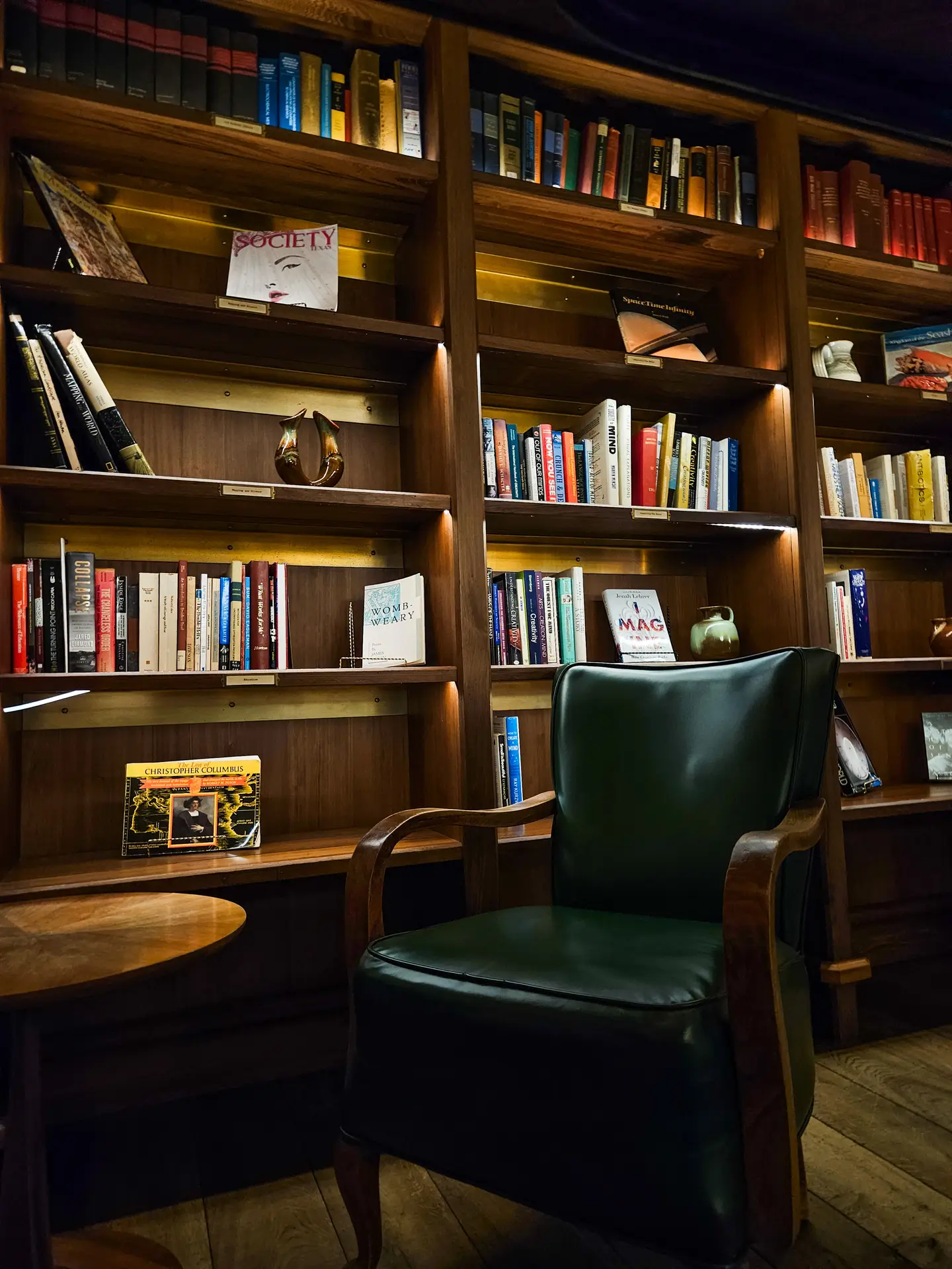 A warm reading nook styled for cozy living rooms, featuring bookshelves with lighting that illuminate rows of colorful books and decorative objects. In front of the shelves stands a dark green leather armchair with wooden armrests, creating an inviting spot to unwind in a softly lit space.