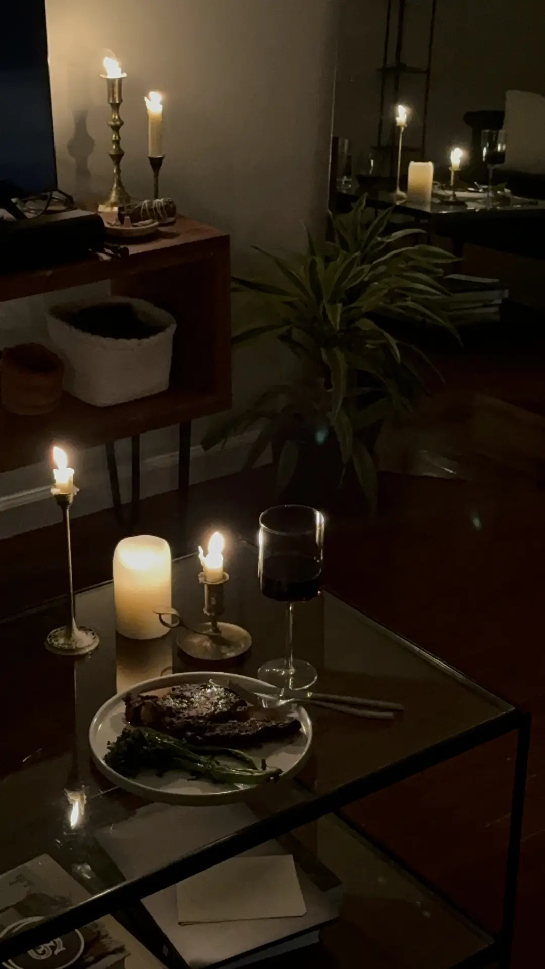 A warm, dimly lit scene in cozy living rooms featuring candles everywhere. Several glowing candles sit on a glass coffee table beside a glass of red wine and a plate of food, while more candlelight shines from a wooden side table in the background. Soft shadows, a houseplant, and dark furniture create an intimate, relaxed atmosphere.