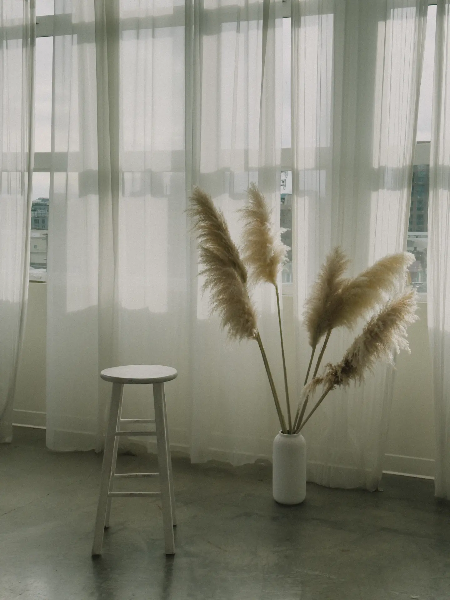 Soft daylight filters through sheer curtains that touch the floor, creating a calm backdrop in cozy living rooms. A simple white stool stands beside a tall vase filled with pampas grass, adding height and airy texture to the minimalist corner.