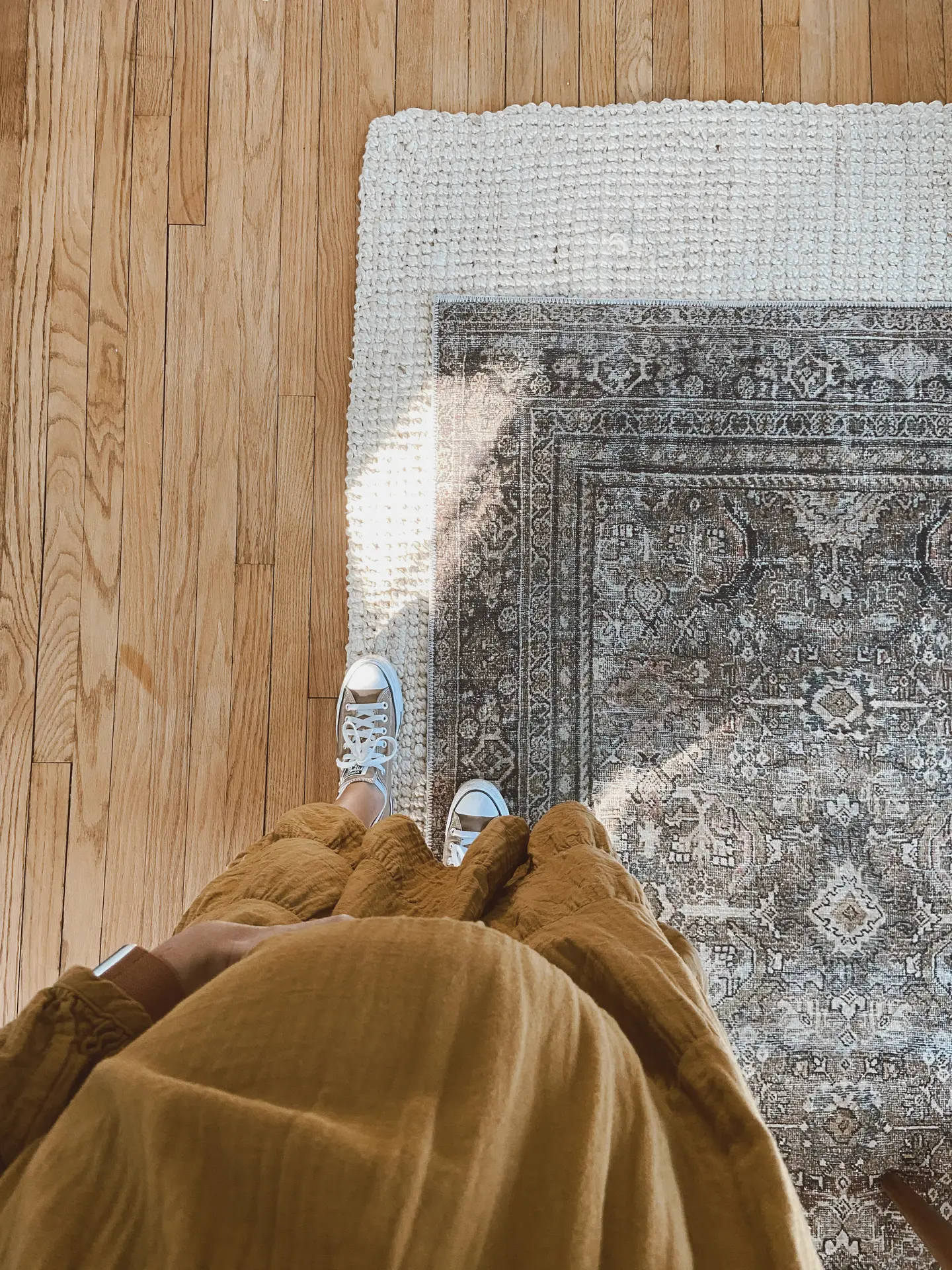 A warm scene showing someone standing in a cozy living room, looking down at layered rugs, a chunky cream woven rug beneath a patterned vintage-style one, placed on natural wood flooring. The outfit's earthy tones blend with the calm palette, creating a relaxed atmosphere perfect for cozy living rooms.