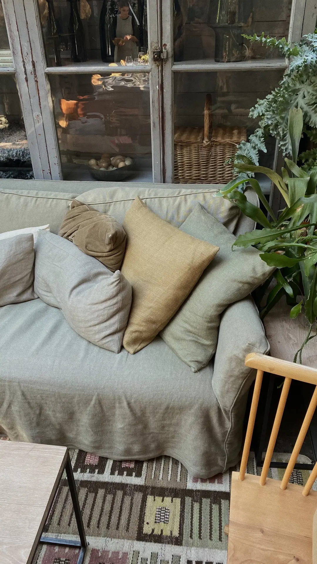 A neutral sofa in a cozy living rooms setting, styled with an assortment of soft, earthy-toned throw pillows. A patterned rug, wooden chair, and leafy green plant add warmth, while a vintage glass cabinet in the background brings character to the relaxed, lived-in space.