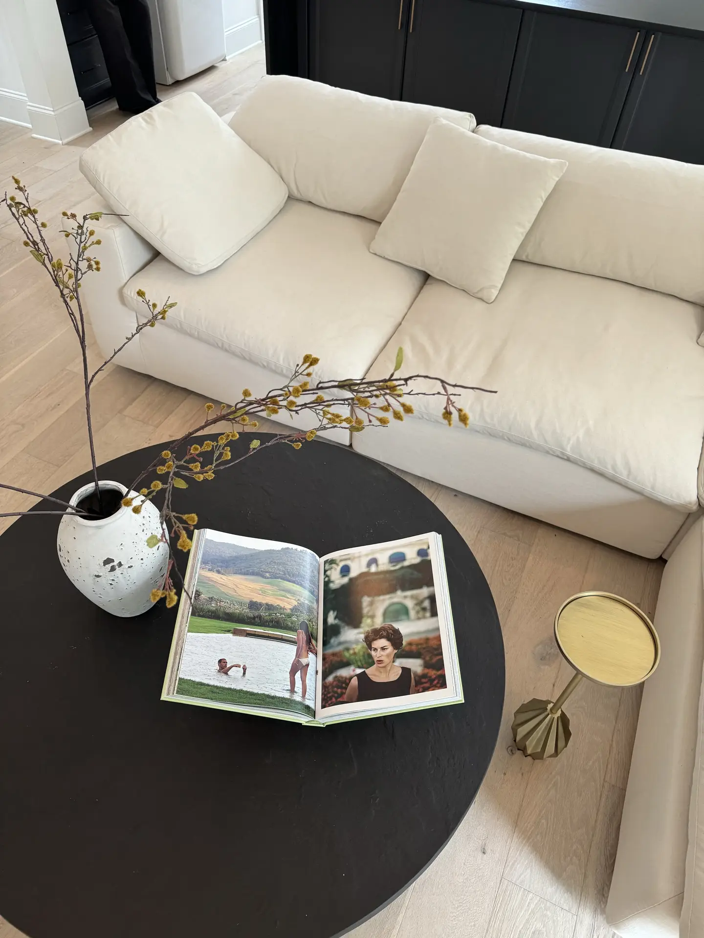 Cozy living rooms often feature simple, inviting setups like this one, where a round coffee table holds an open art book and a ceramic vase with long branches, placed in front of a soft, cream-colored sofa on light wood flooring.