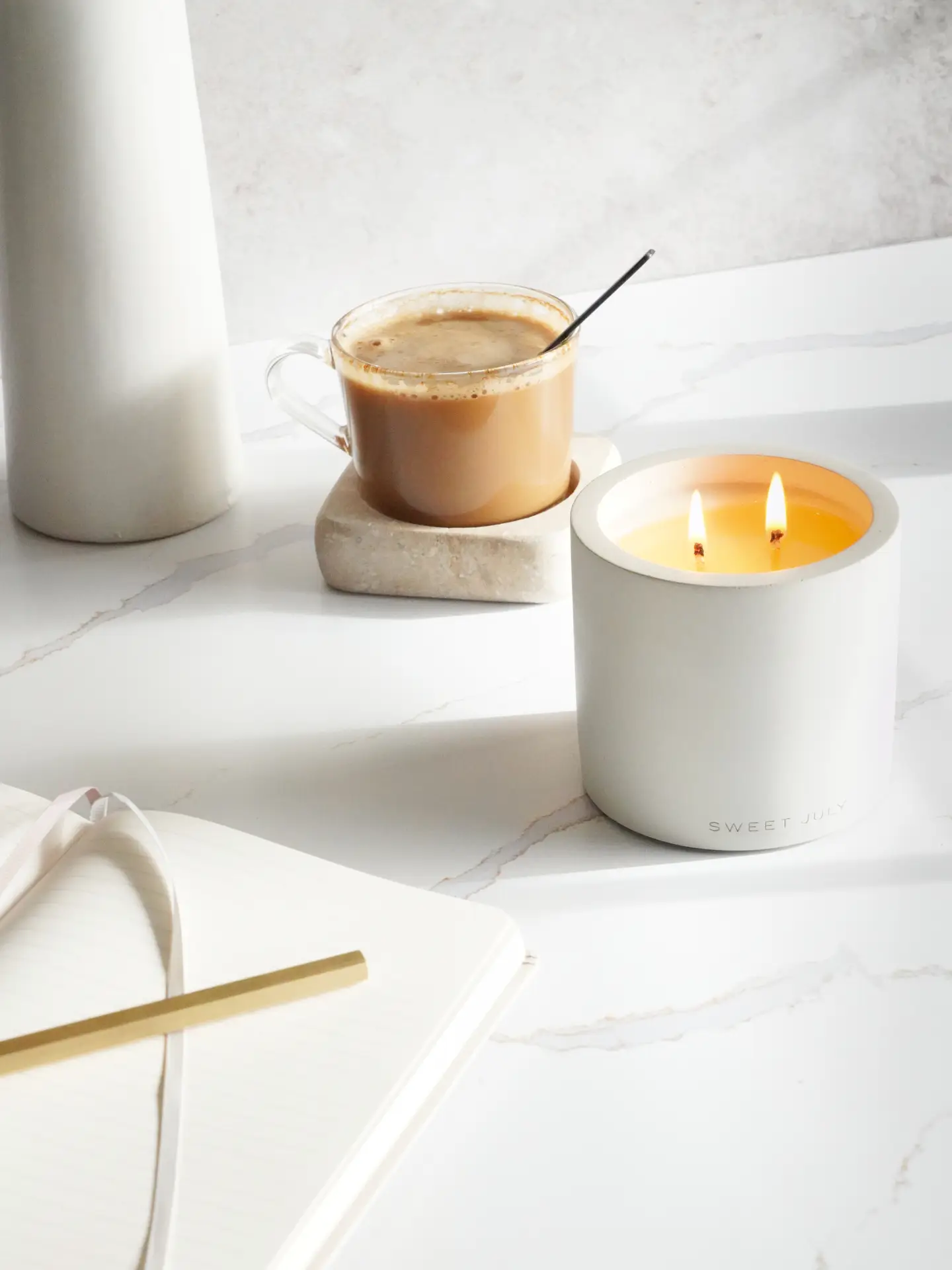 A bright, minimal setup inspired by cozy living rooms, featuring a steaming latte in a clear mug on a stone coaster beside a softly glowing candle that adds subtle scented touches. An open notebook with a gold pen rests on a clean white surface, creating a calm and inviting moment for journaling or unwinding.