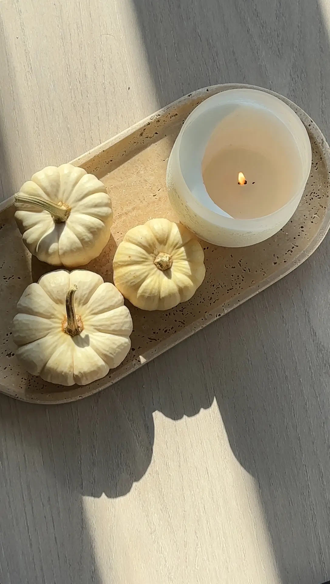 A close-up of a light-toned tray on a wooden surface, holding three small white pumpkins and a softly glowing candle, lit by warm sunlight. The scene hints at cozy living rooms and a seasonal decor rotation, creating a gentle autumn atmosphere through simple, natural accents.