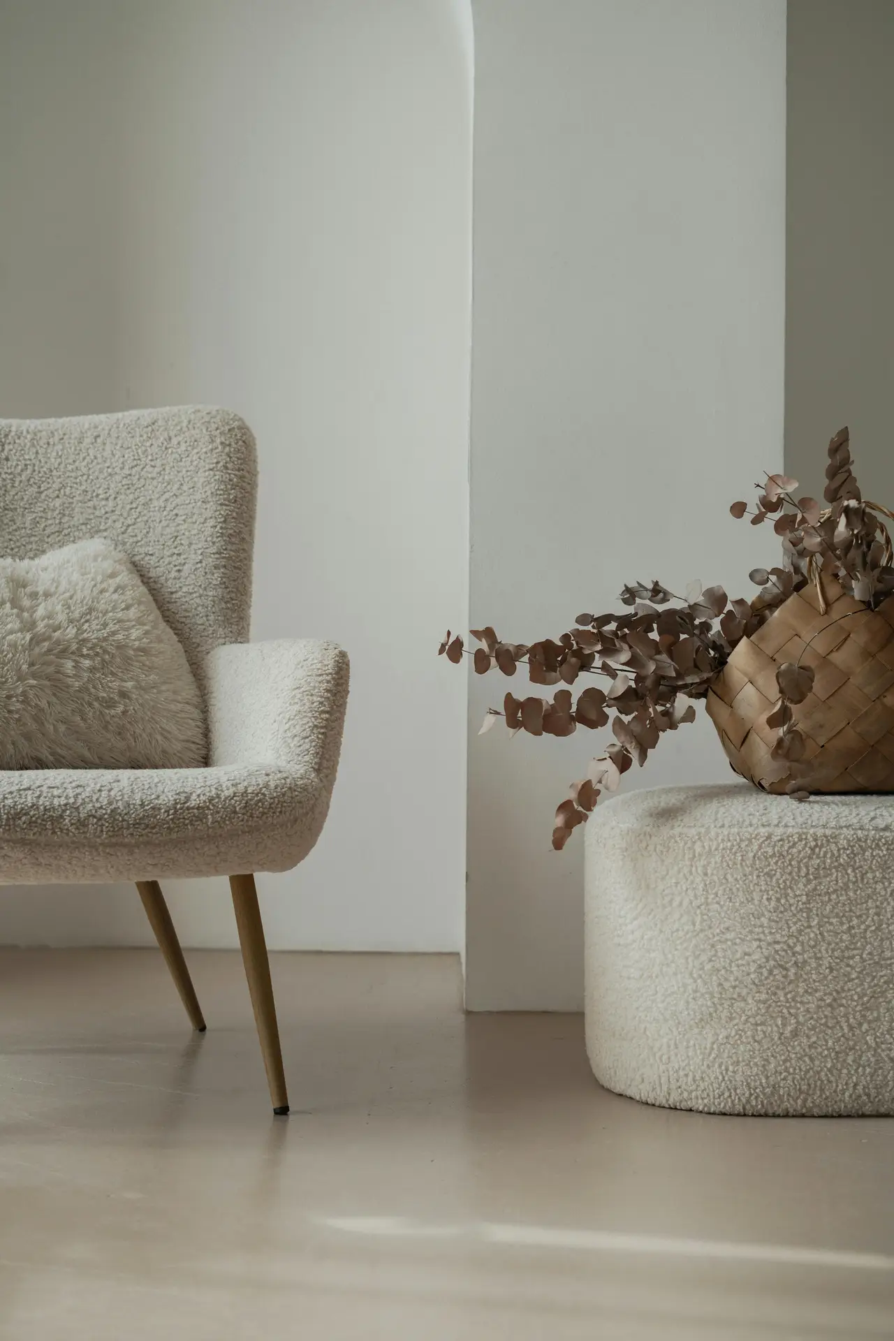 A serene corner often seen in cozy living rooms, featuring a boucle armchair with a plush pillow and a matching upholstered ottoman. A woven basket filled with dried eucalyptus adds warmth, while the mix of soft textures and neutral tones creates a calm, inviting atmosphere against the simple backdrop.