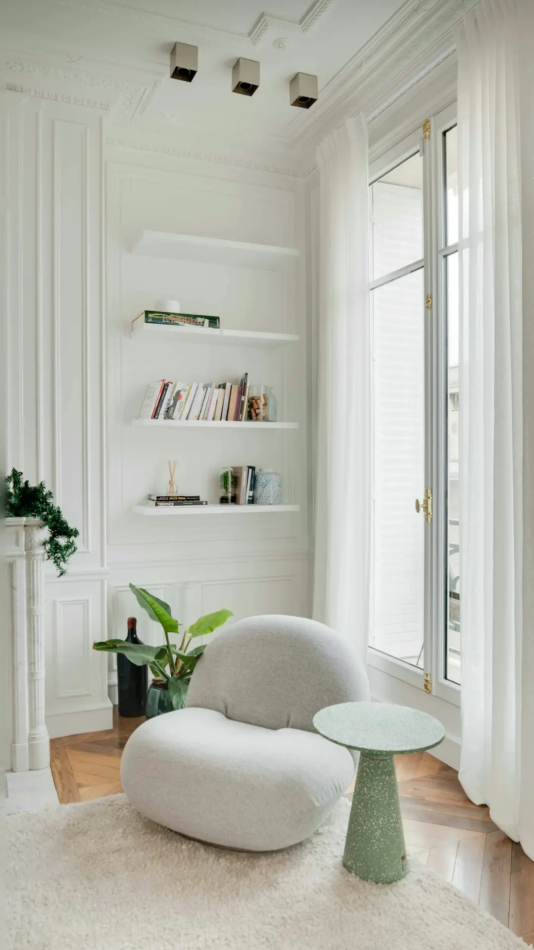 A bright corner of a cozy living room featuring a soft, rounded statement armchair in light gray, paired with a small green side table on a plush rug. Built-in white shelves filled with books and decorative accents line the wall beside tall windows framed with airy curtains, creating an inviting and serene atmosphere.