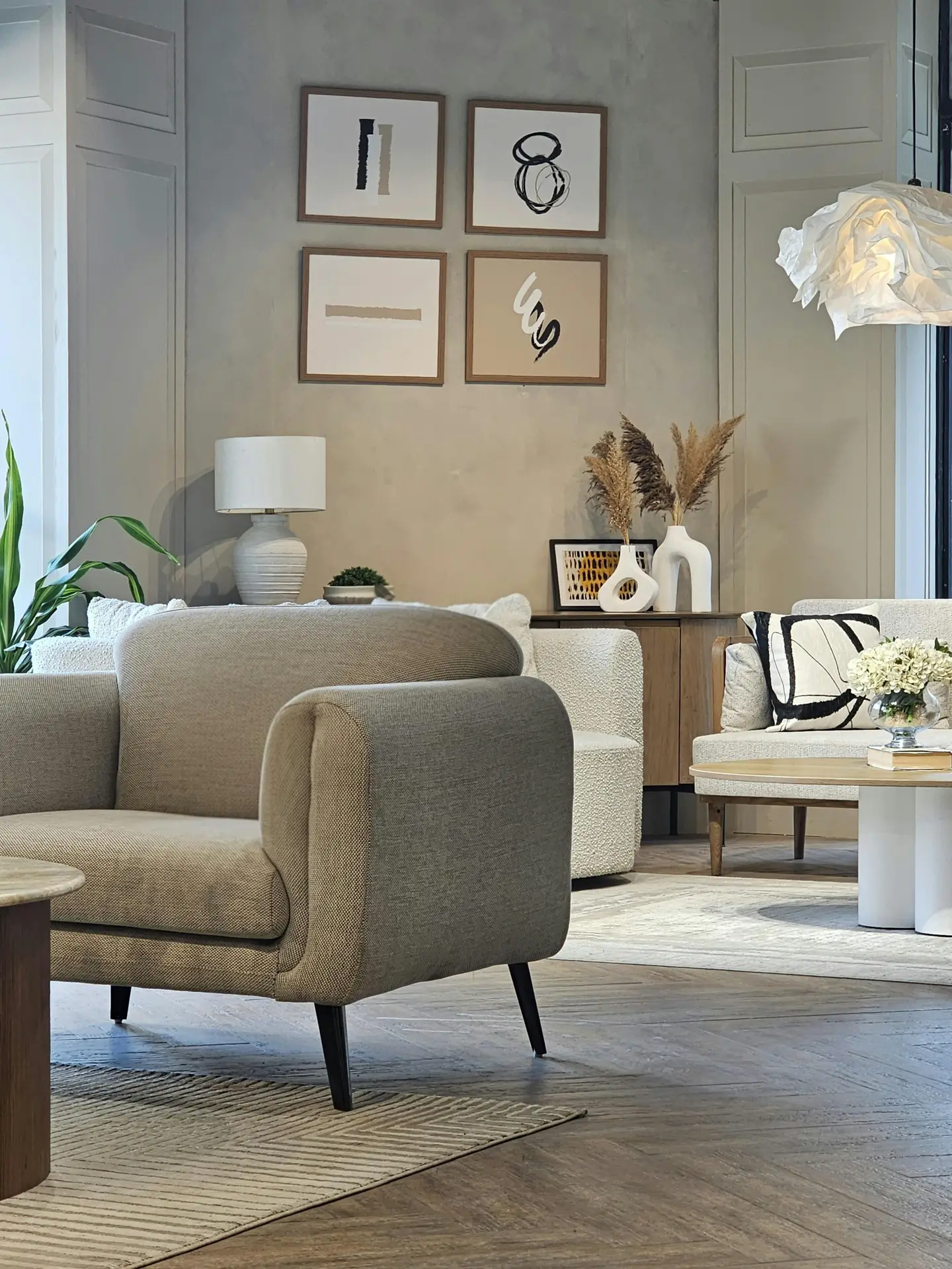 A stylish, cozy living room arranged in a tonal monochrome palette, featuring a soft beige sofa in the foreground, layered neutral rugs, and abstract framed art on the wall. In the background, a white accent chair, pampas grass in sculptural vases, and warm wood furniture create a calm, cohesive atmosphere.