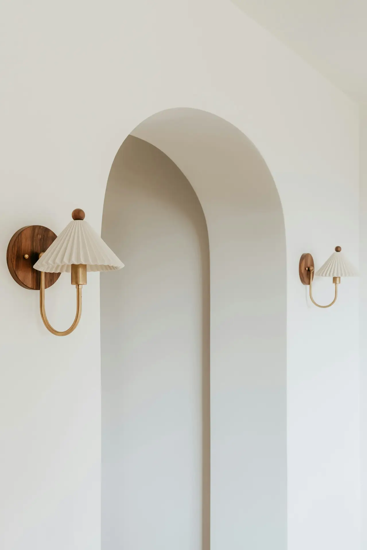 A pair of warm ambient wall sconces mounted on either side of a soft, curved archway, adding gentle light and architectural interest to a cozy living room. The minimalist design and neutral tones create a calm, versatile atmosphere.