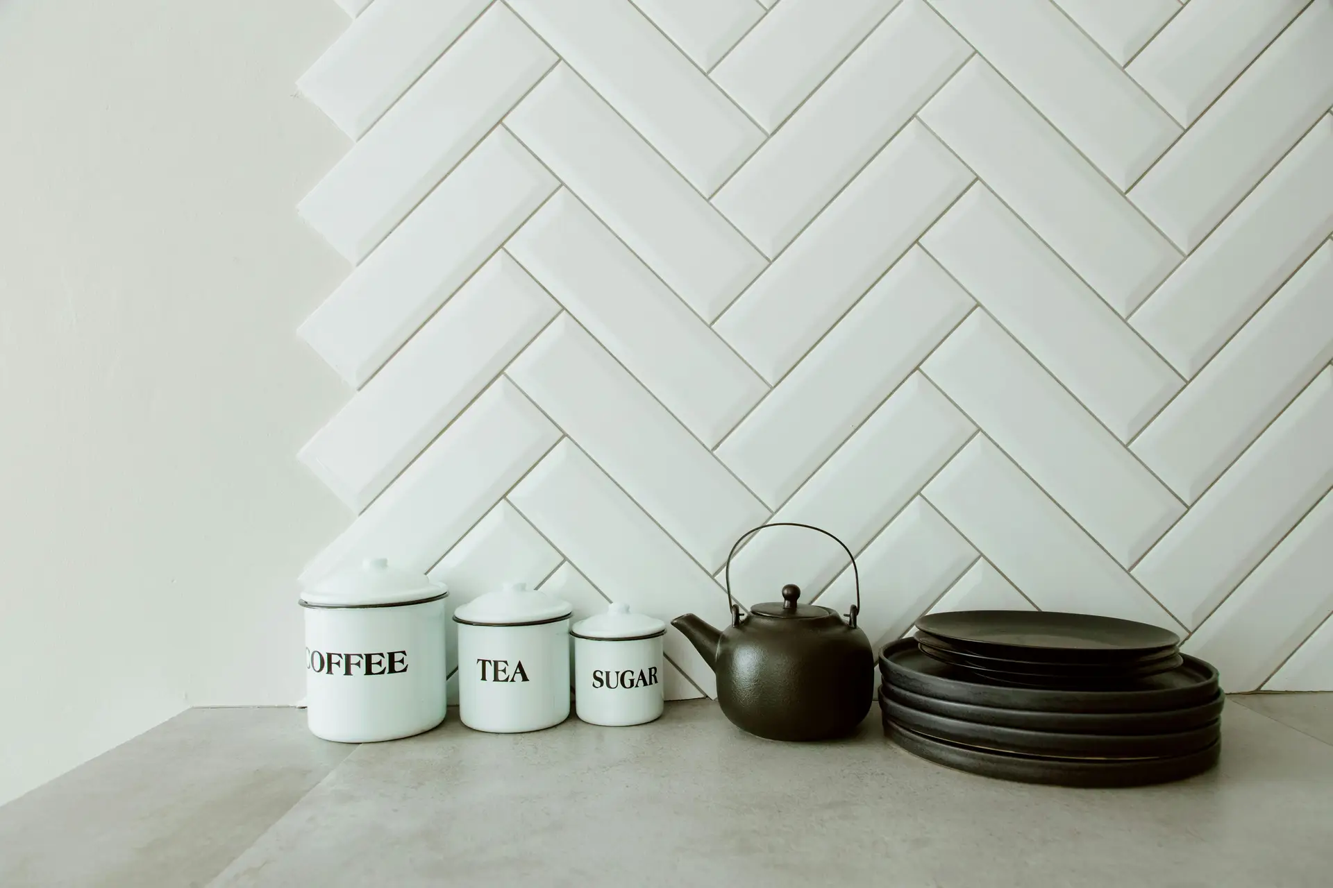 A minimalist kitchen counter featuring white canisters labeled coffee, tea, and sugar, a black teapot, and stacked dark plates set against a wall of beveled subway tiles in a herringbone pattern. The clean lines and soft contrast provide modern kitchen backsplash ideas that blend simplicity with subtle texture.