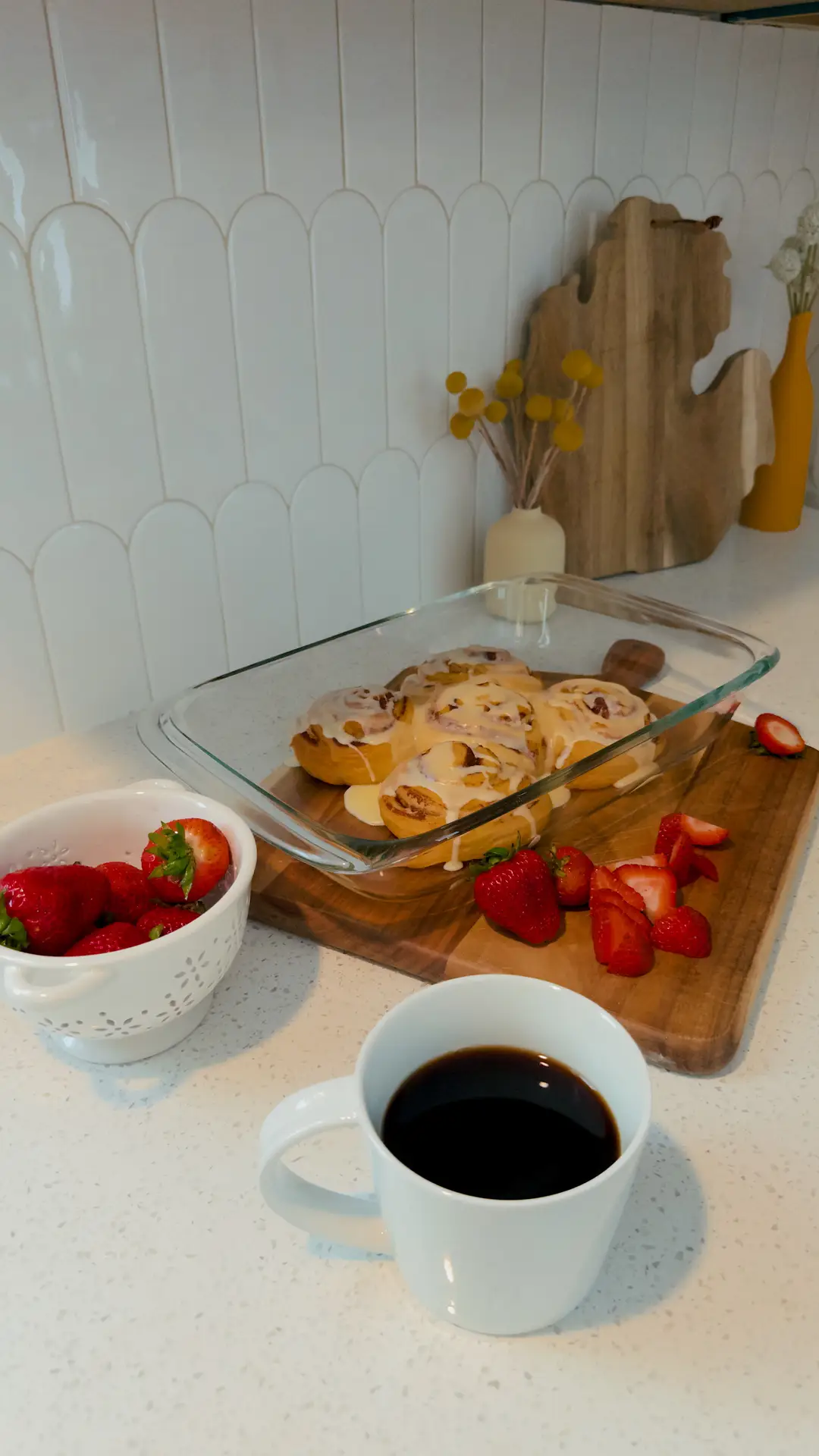 A bright kitchen countertop featuring a tray of cinnamon rolls, fresh strawberries, and a cup of coffee set against a glossy white backsplash made of fish scale tiles. The curved tile shape adds a soft, modern touch, offering fresh kitchen backsplash ideas that balance texture and simplicity for everyday spaces.
