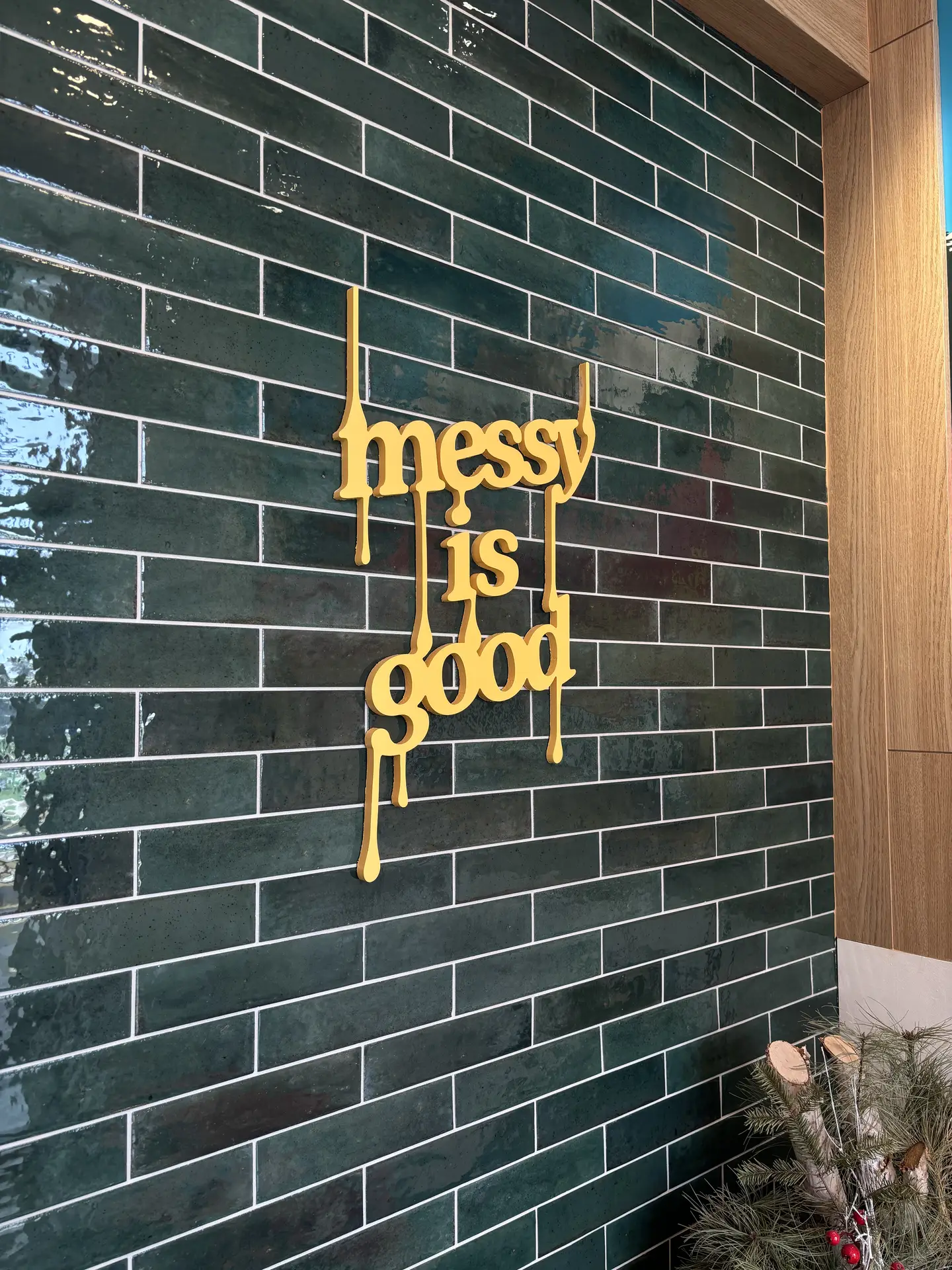 A wall covered in glossy green subway tiles framed by light wood panels, featuring a yellow decorative sign that reads “messy is good.” The rich color and reflective finish of the tiles add depth and character, offering creative kitchen backsplash ideas for modern or eclectic interiors.