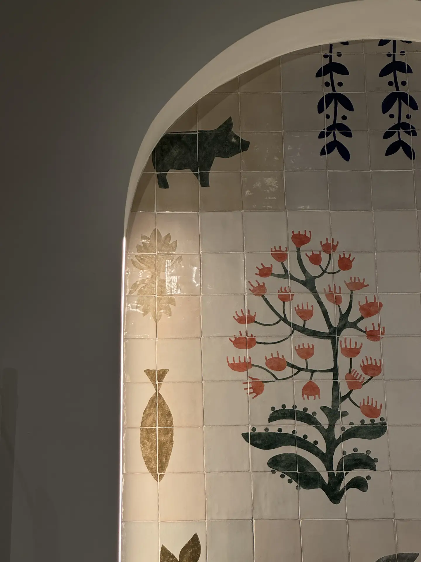 An arched wall section decorated with hand-painted wall tiles featuring whimsical designs like a tree with red flowers, a pig, and abstract plant shapes. The soft lighting highlights the artisanal glaze and texture, making it a creative example of kitchen backsplash ideas that blend craftsmanship and artistic charm.