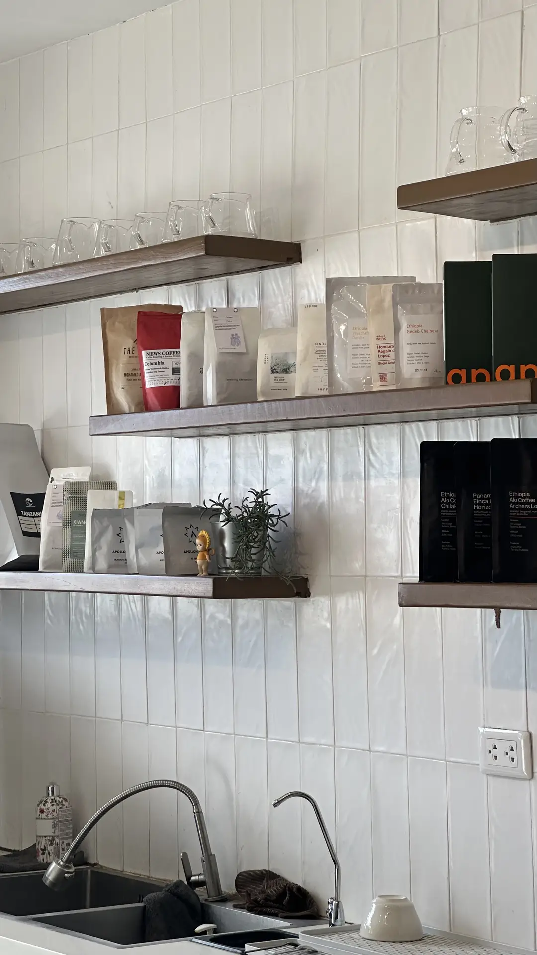 A modern kitchen setup with open wooden shelves displaying coffee bags and glassware, paired with a glossy white backsplash made of vertical kit kat tiles. The slim, elongated tile design adds a subtle texture and contemporary flair, making it a stylish example of kitchen backsplash ideas for minimalist or café-inspired interiors.