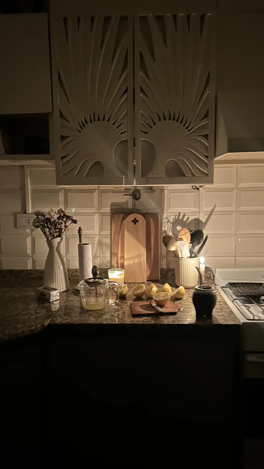 A softly lit kitchen scene featuring a countertop with lemons, utensils, and a lit candle, creating a warm and cozy atmosphere. The wall behind the counter showcases raised panel subway tiles that add depth and character to the space, making it an inspiring example for kitchen backsplash ideas that balance classic texture with inviting charm.