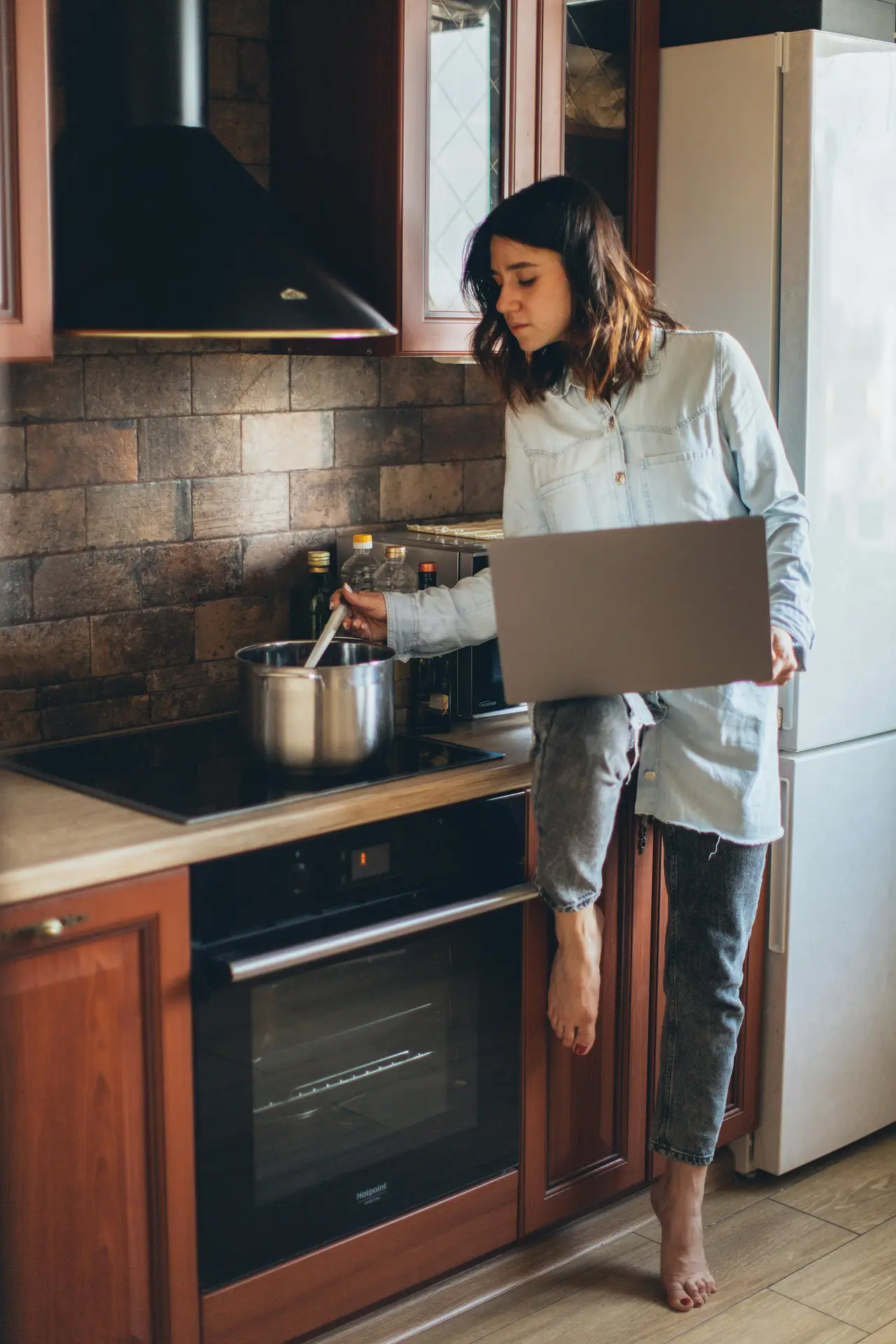 A cozy kitchen with wooden cabinets and a backsplash made of rustic stone tiles, creating a warm and textured backdrop. A woman in casual clothes stirs a pot on the stove while checking something on her laptop, blending everyday life with inviting kitchen backsplash ideas for traditional or farmhouse-style spaces.