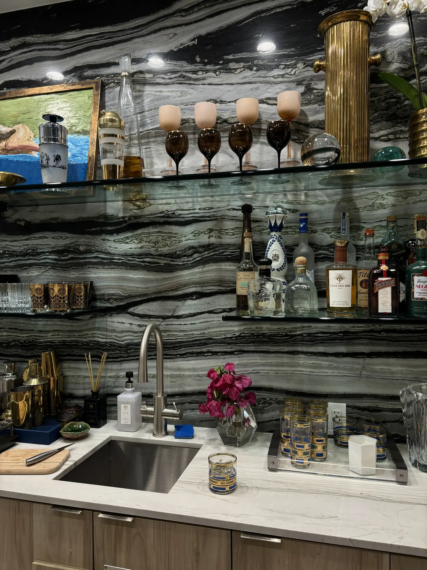 A stylish bar-style kitchen setup featuring a striking striped marble slab backsplash that adds depth and luxury to the space. Glass shelves hold elegant drinkware, bottles, and decorative pieces, while the countertop displays a mix of gold accents and fresh flowers. This setting presents kitchen backsplash ideas that merge sophistication with bold natural stone textures.