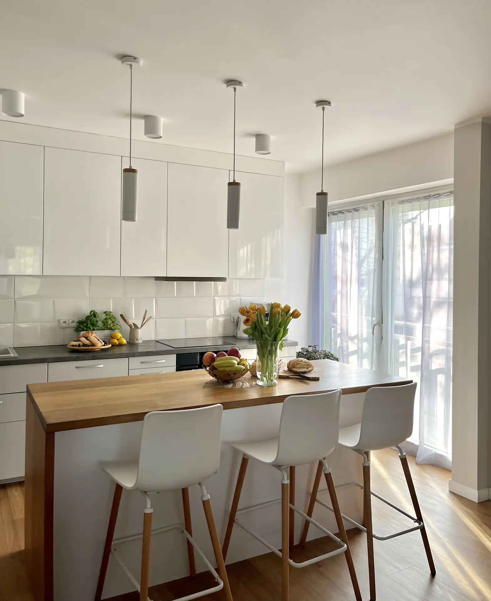 A bright contemporary kitchen with a wooden island, white cabinetry, and three pendant lights hanging above. The glossy subway tiles on the backsplash add texture and reflect natural light from the nearby window, offering simple yet elegant kitchen backsplash ideas for modern, minimalist spaces.