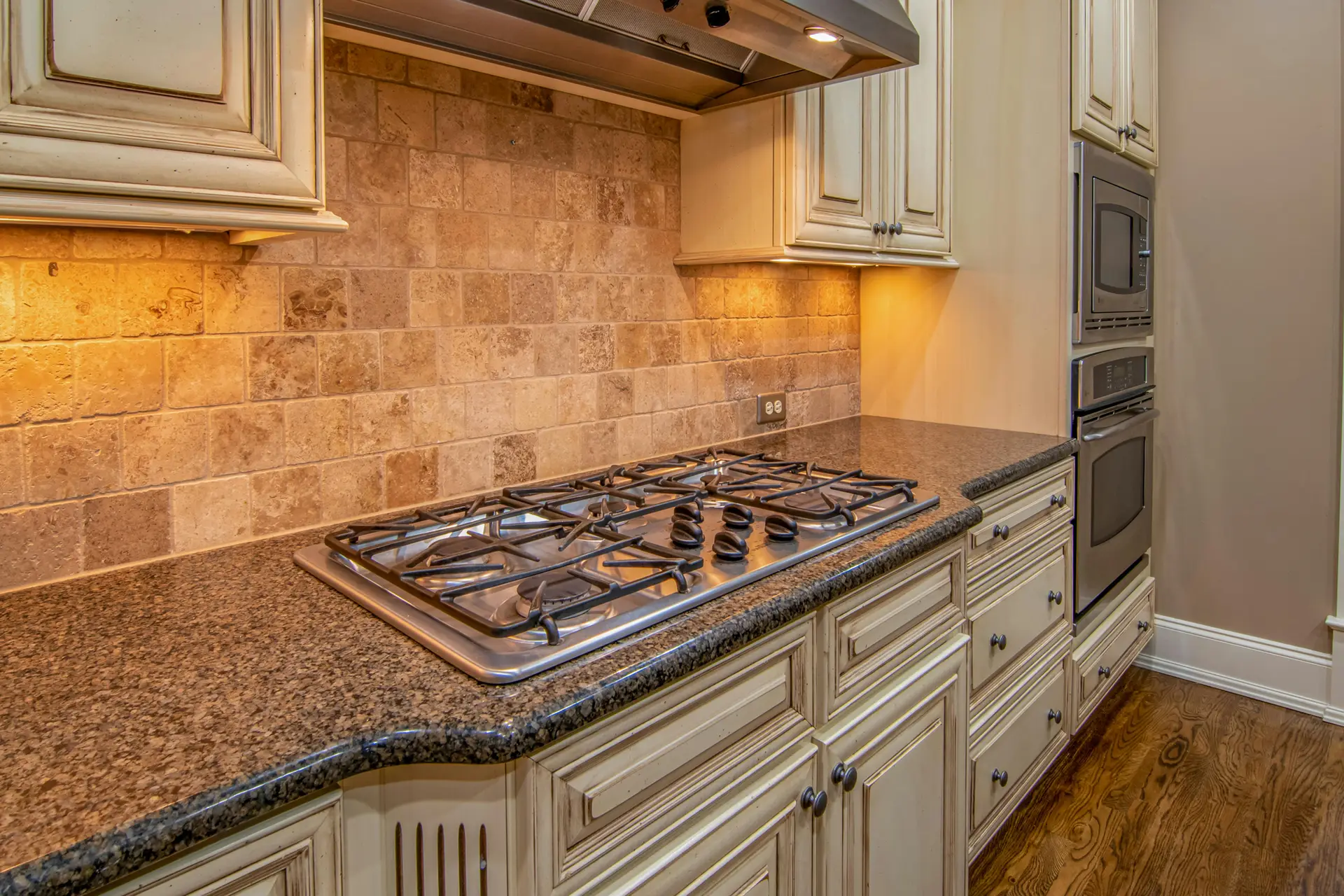 A traditional kitchen design featuring a backsplash made of tumbled travertine tiles in warm beige and tan tones, adding natural texture and depth. The space includes cream cabinetry, granite countertops, and a stainless steel cooktop, showcasing kitchen backsplash ideas that bring timeless charm and earthy elegance to classic interiors.