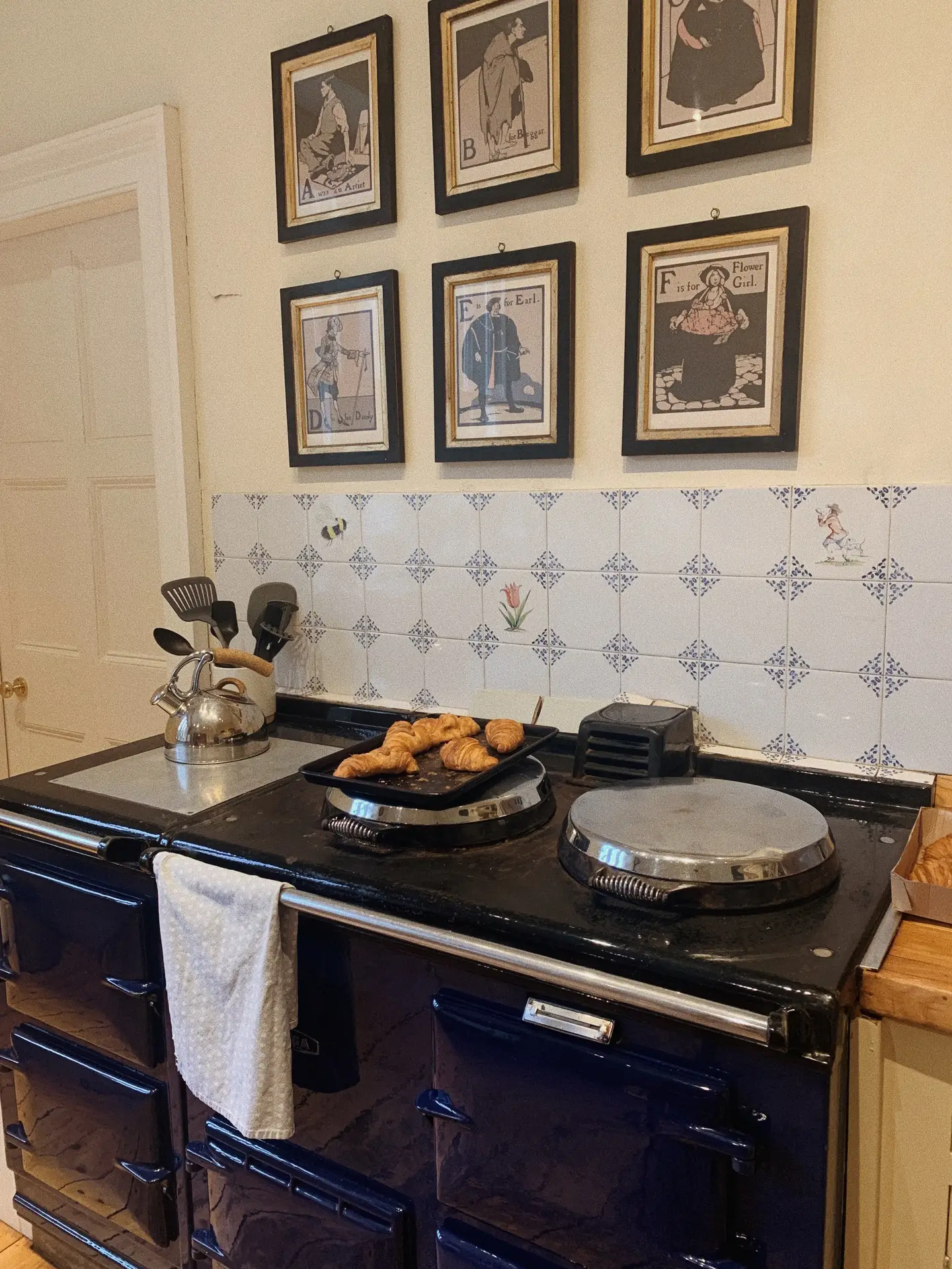 A cozy traditional kitchen featuring a dark enamel stove with freshly baked croissants, framed by a backsplash of vintage delft tiles. The delicate blue floral and figurative motifs add nostalgic charm, offering timeless kitchen backsplash ideas that blend classic craftsmanship with a warm, lived-in feel.