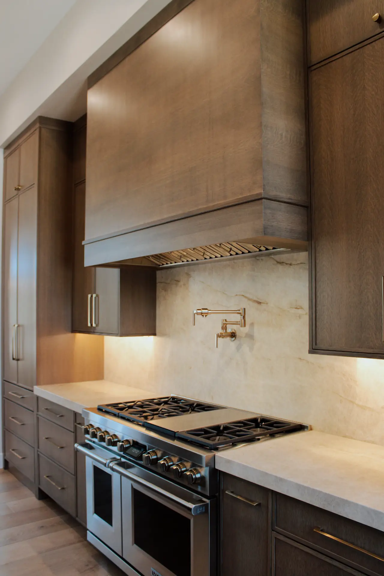 A contemporary kitchen featuring dark wood cabinetry, brass fixtures, and a sleek white marble slab backsplash behind a stainless steel range. The seamless stone surface adds luxury and sophistication, providing elegant kitchen backsplash ideas for modern homes that favor clean lines and natural materials.