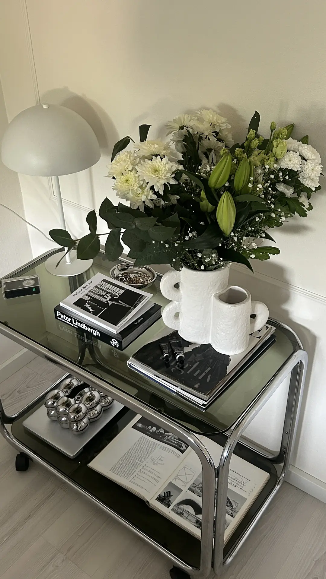 A stylish living room corner featuring a chrome and glass side table styled with fresh white flowers, coffee table books, and a sculptural lamp. This setup demonstrates living room ideas for corner maximization by turning a small space into a curated display that blends functionality with modern elegance.
