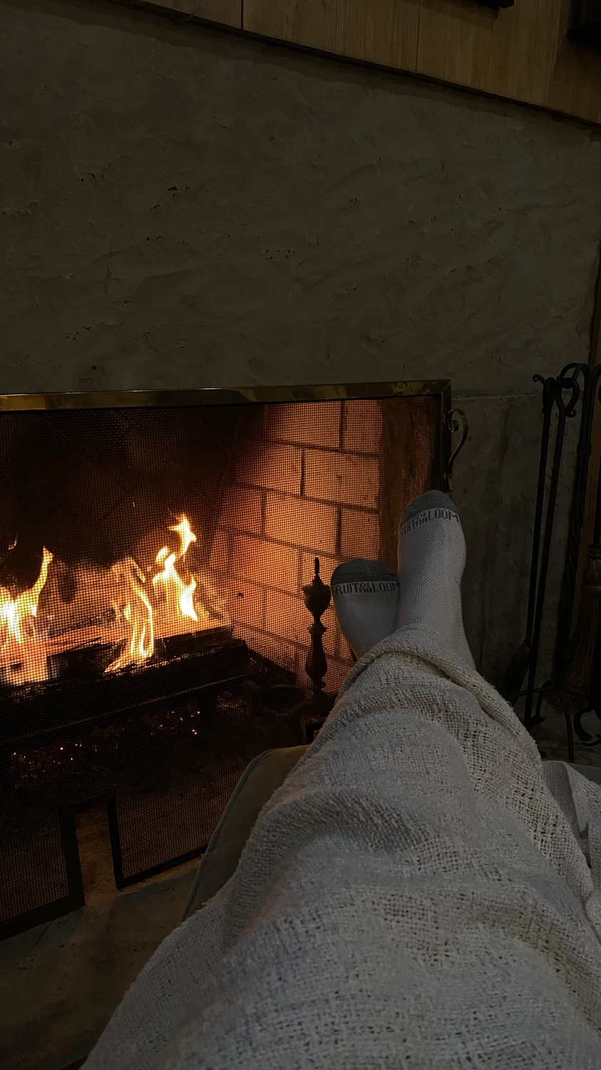 A warm living room scene with a person relaxing by a glowing fire, feet wrapped in a blanket and propped up toward the hearth. The soft lighting and textured materials showcase living room ideas that focus on a cozy fireplace (reimagined) for calm, intimate evenings.