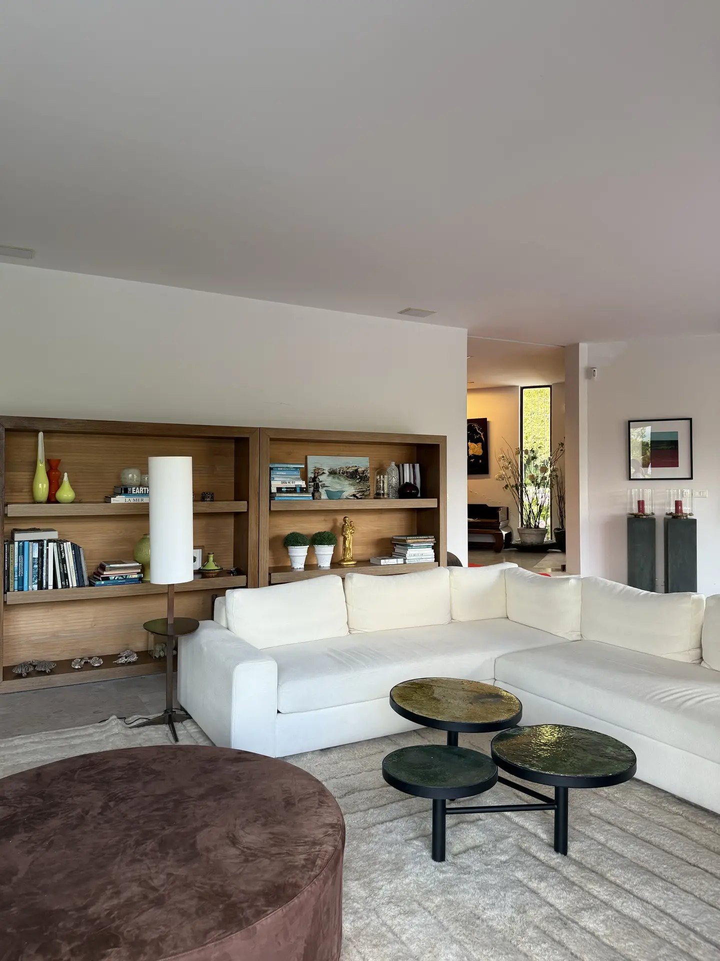 A contemporary living room with a floating furniture arrangement featuring a large white sectional sofa centered on a neutral rug. The space includes layered coffee tables with round tops, a brown ottoman, and open wooden shelving filled with books and decorative objects. This layout highlights living room ideas that create openness and balance while maintaining a cozy, welcoming atmosphere.