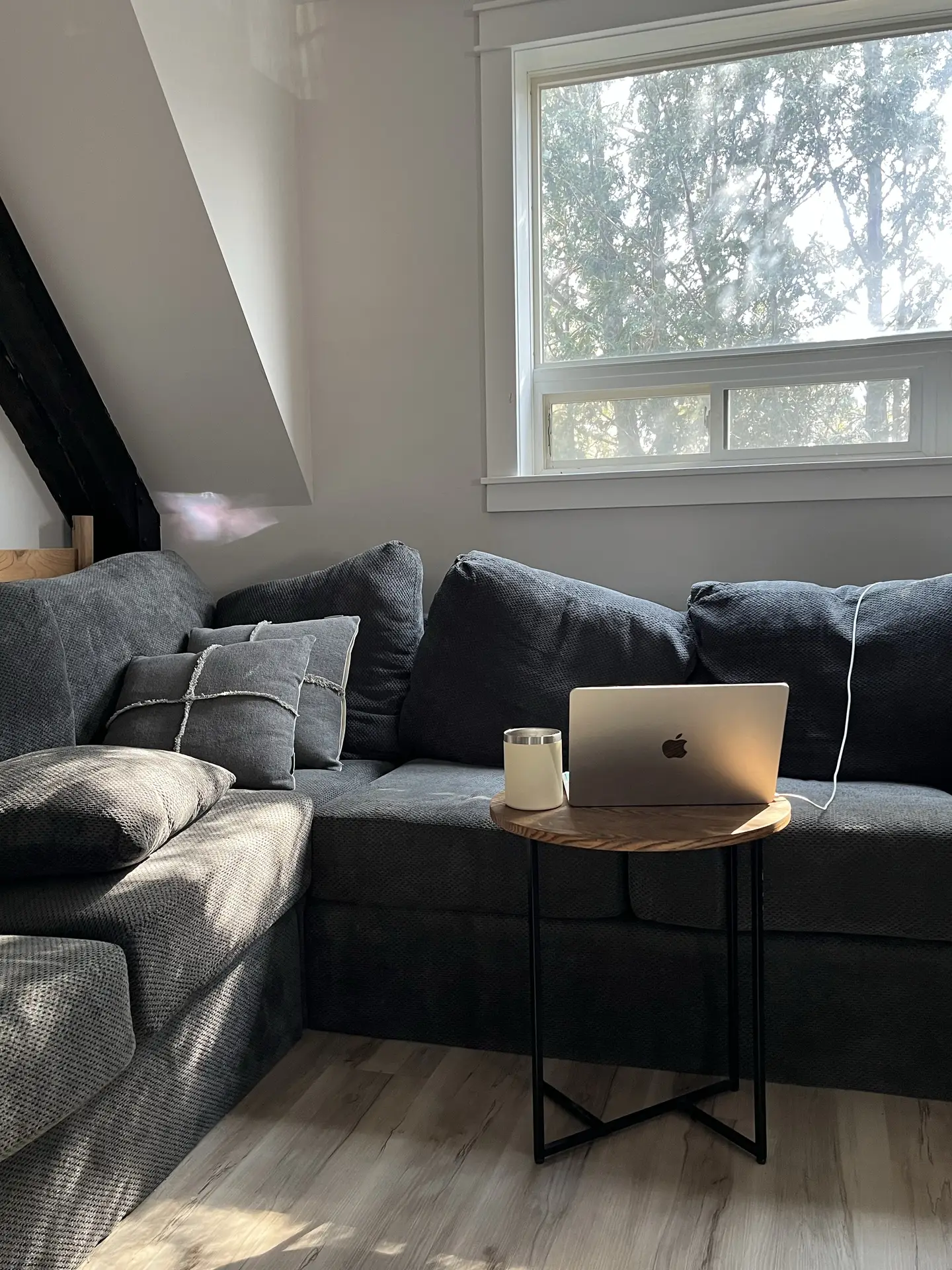 A bright corner living room featuring a dark gray L-shaped sectional with textured fabric, paired with a small round wooden side table holding a laptop and candle. This setup captures living room ideas focused on cozy efficiency and relaxed, multi-purpose lounging spaces.