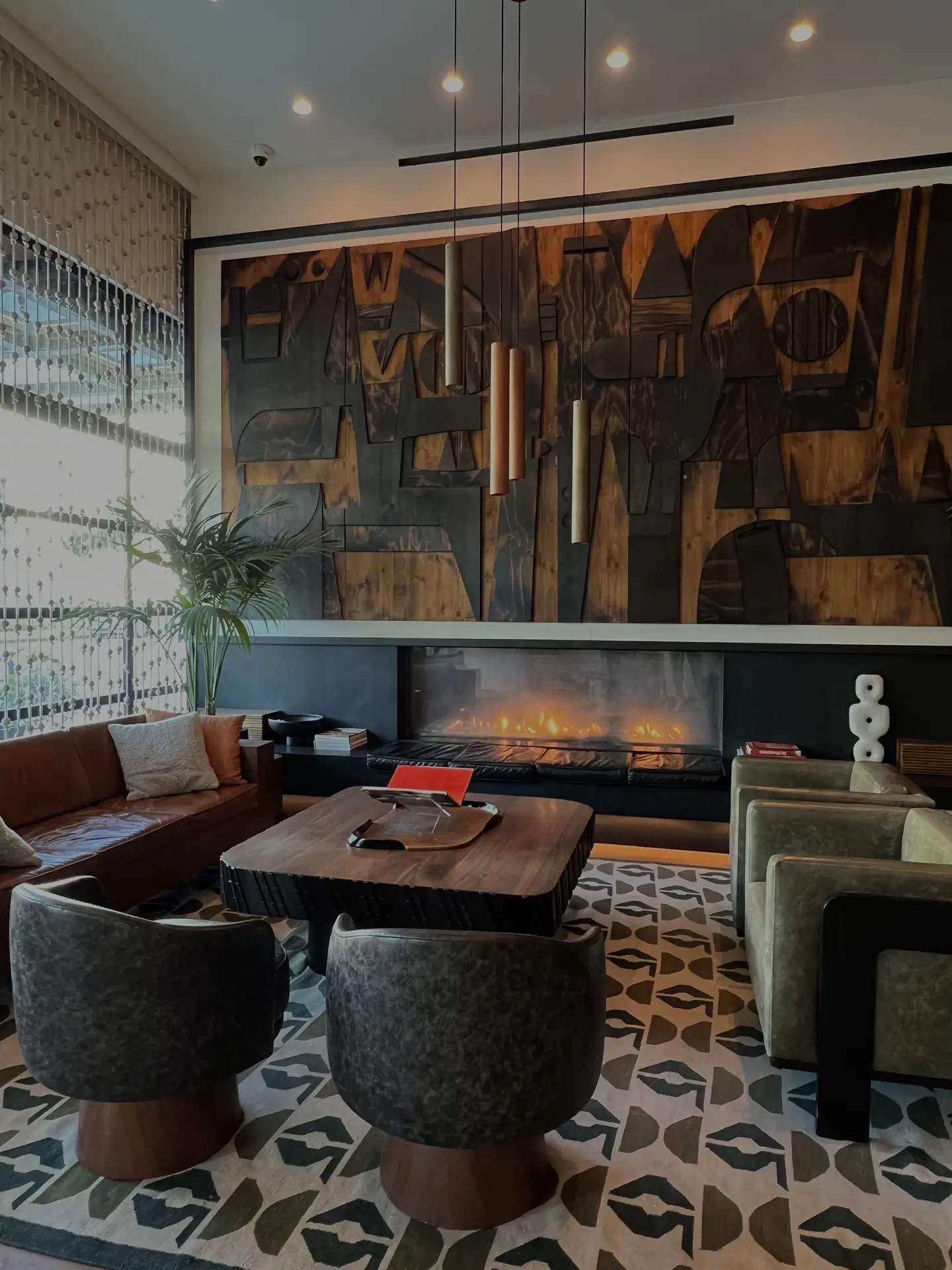 A sophisticated living room featuring a mix of modern and mid-century design with a geometric rug, leather and velvet seating, and a wooden coffee table centered by a glowing fireplace. Pendant lights and recessed ceiling spots create layered lighting, adding depth and warmth to the earthy tones and textured artwork on the wall.