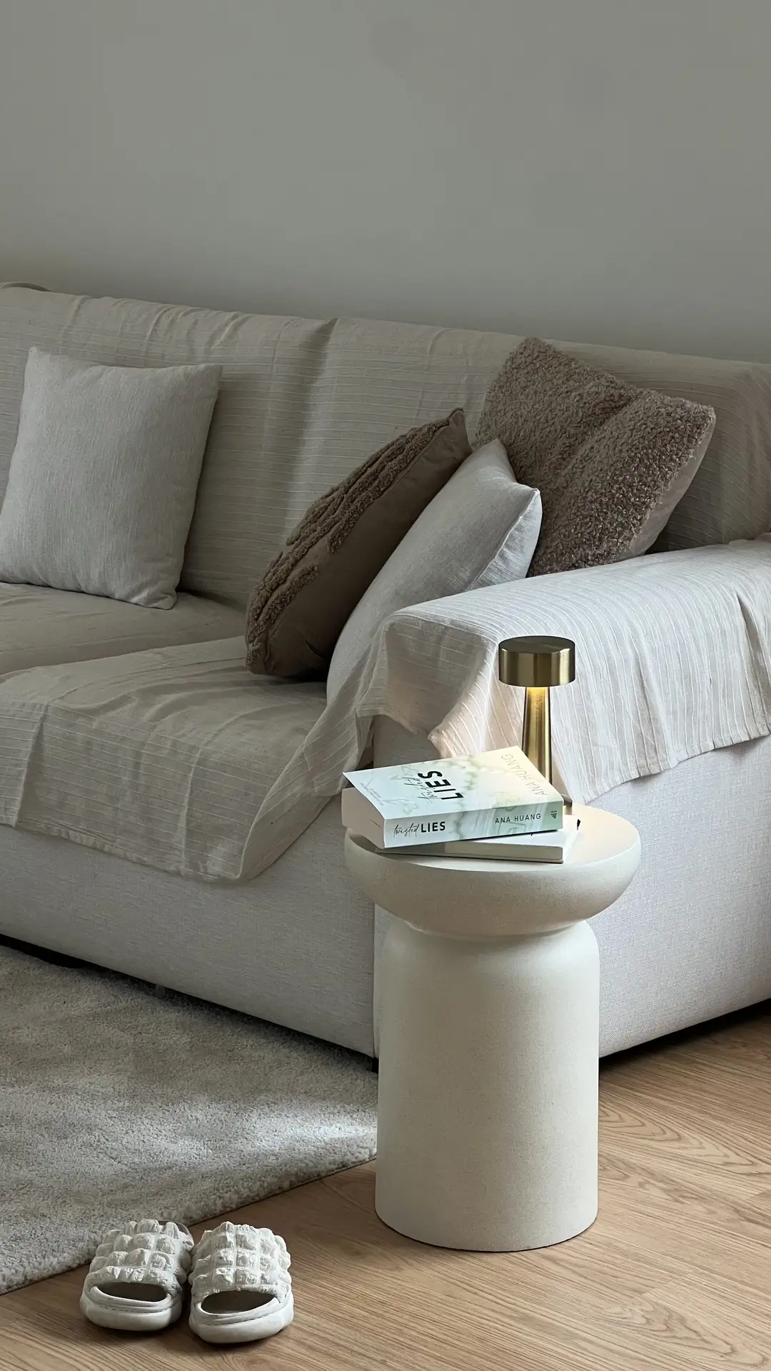 A minimalist living room styled in soft neutral tones, featuring a beige sofa with textured throw pillows and a small sculptural side table holding books and a brass lamp. A cozy rug and slippers add warmth to the space, reflecting living room ideas that focus on monochromatic layering for a calm and cohesive aesthetic.