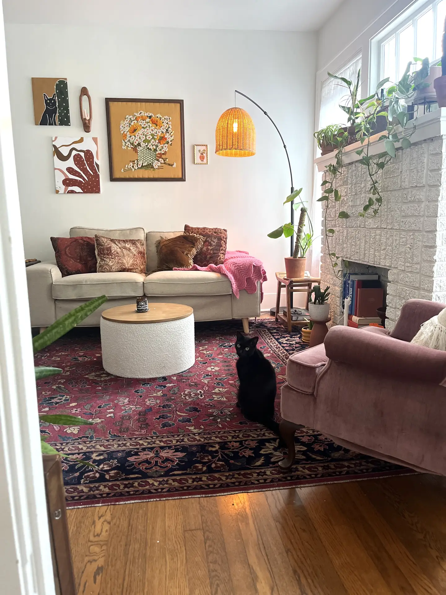 A cozy living room with a mix of vintage and modern touches, featuring a beige sofa with patterned cushions, a pink velvet armchair, and a round white coffee table on a red oriental rug. Plants on the mantel and artwork on the wall add warmth and personality, showcasing living room ideas that create multi-functional zones for relaxation, reading, and conversation.