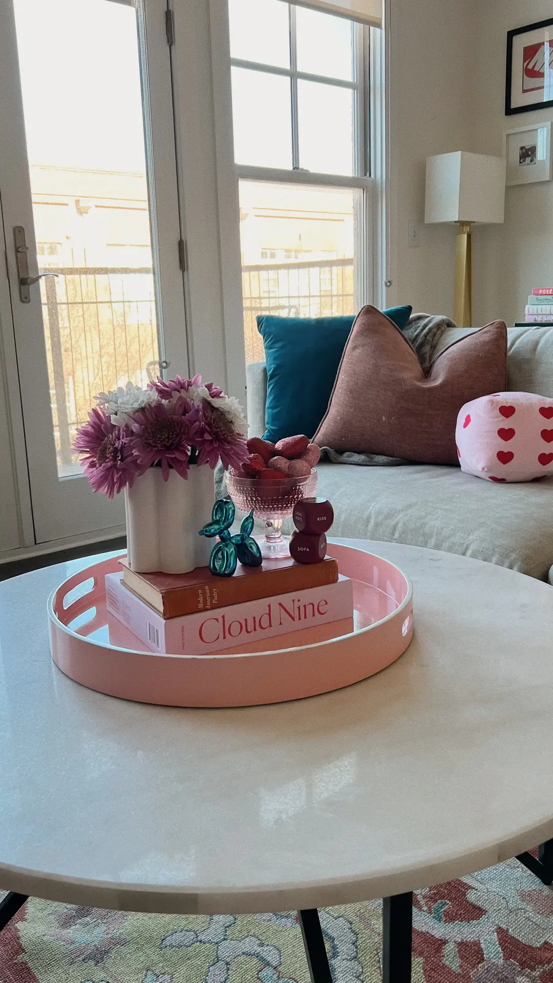A bright living room with a neutral sofa styled with colorful throw pillows in teal and blush tones, complemented by a round coffee table featuring a pink tray, flowers, and decor pieces. This setup showcases living room ideas using a natural base and bold accents to create a cheerful, modern vibe.