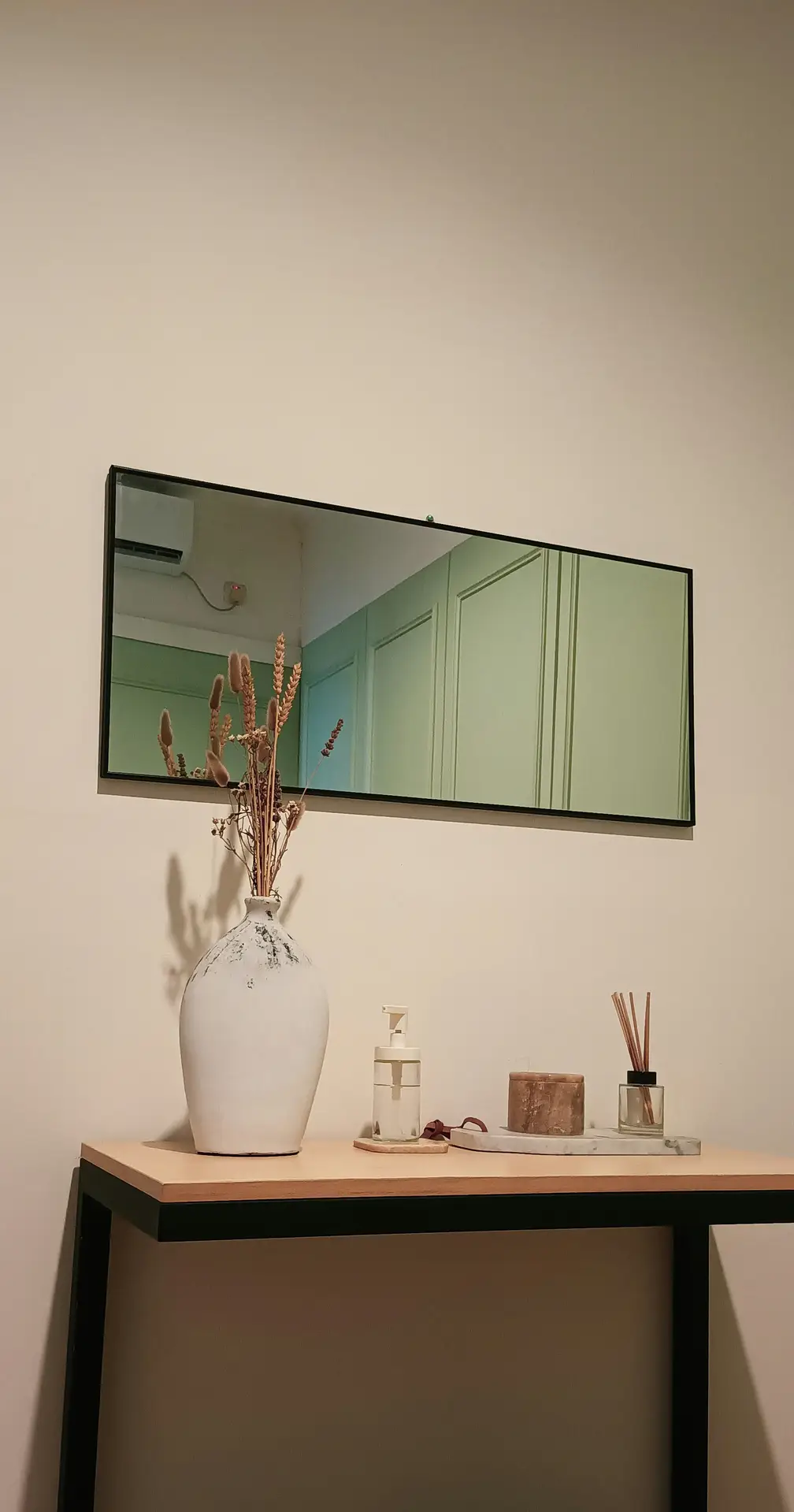 A minimal living room setup with a slim console table decorated with a ceramic vase of dried flowers, a candle, and a reed diffuser beneath a rectangular mirror. This image captures living room ideas that use reflective surfaces to visually expand space and enhance natural light.
