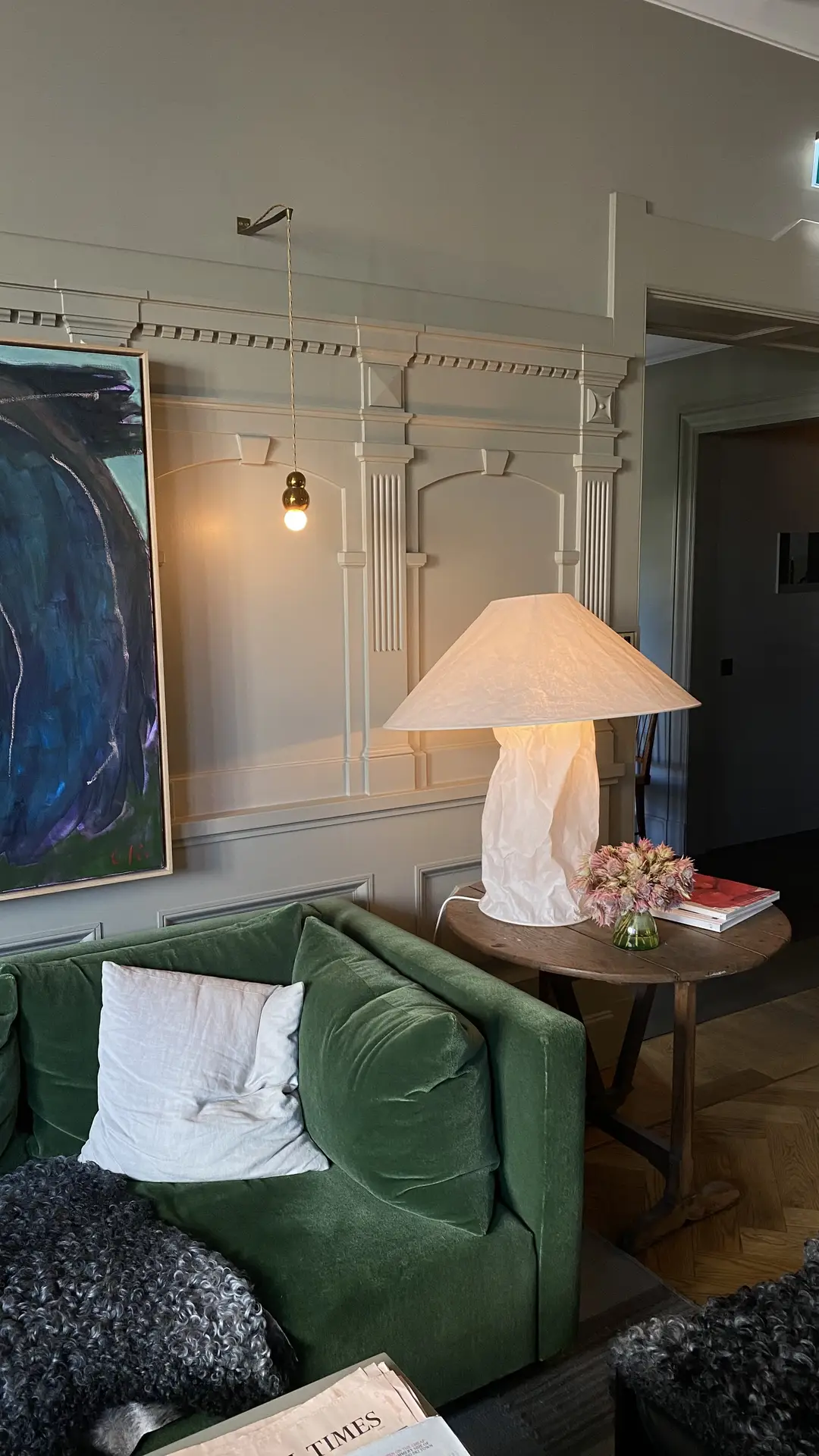 A sophisticated living room featuring a green velvet sofa, neutral walls with decorative moldings, and an eye-catching mix of lighting elements, a sculptural paper table lamp and a hanging bulb with a brass fitting. A bold abstract painting and fresh flowers complete the scene, illustrating living room ideas that use statement lighting as art to enhance character and atmosphere.