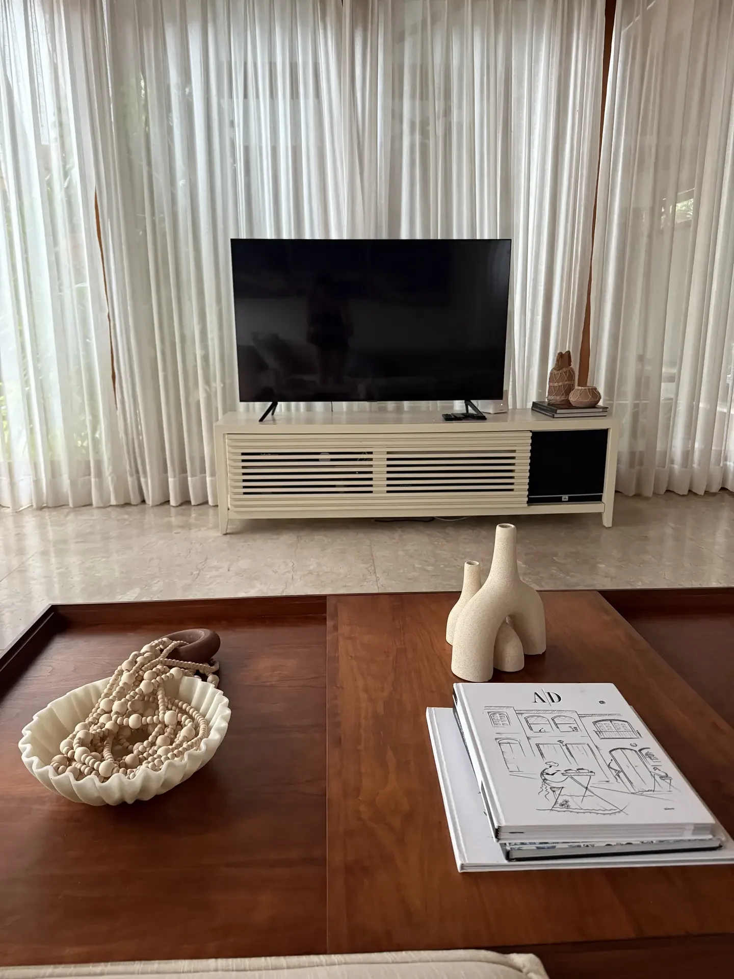 A minimal living room with a sleek TV mounted above a modern slatted console, paired with neutral-toned decor and sculptural accents on a wooden coffee table. This setup reflects living room ideas centered on tech-integrated design that blends functionality with calm, aesthetic balance.
