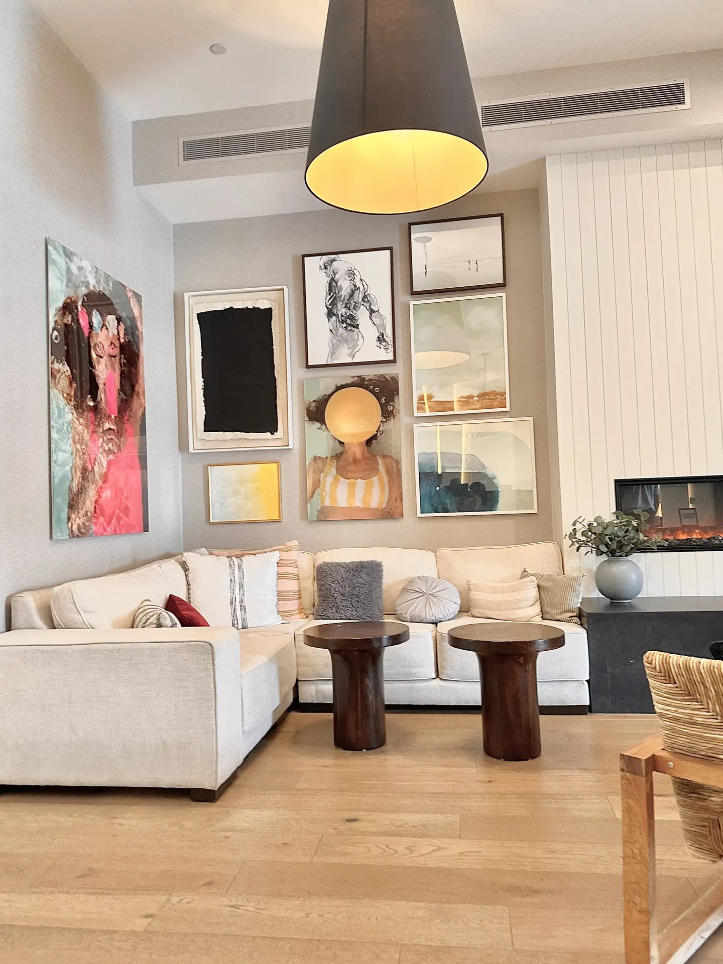 A modern living room with a beige sectional sofa, wooden side tables, and layered throw pillows arranged in a cozy corner. The focal point is a striking accent wall statement featuring a curated gallery of abstract and portrait artwork, illuminated by a large black pendant light that adds warmth and character to the space.