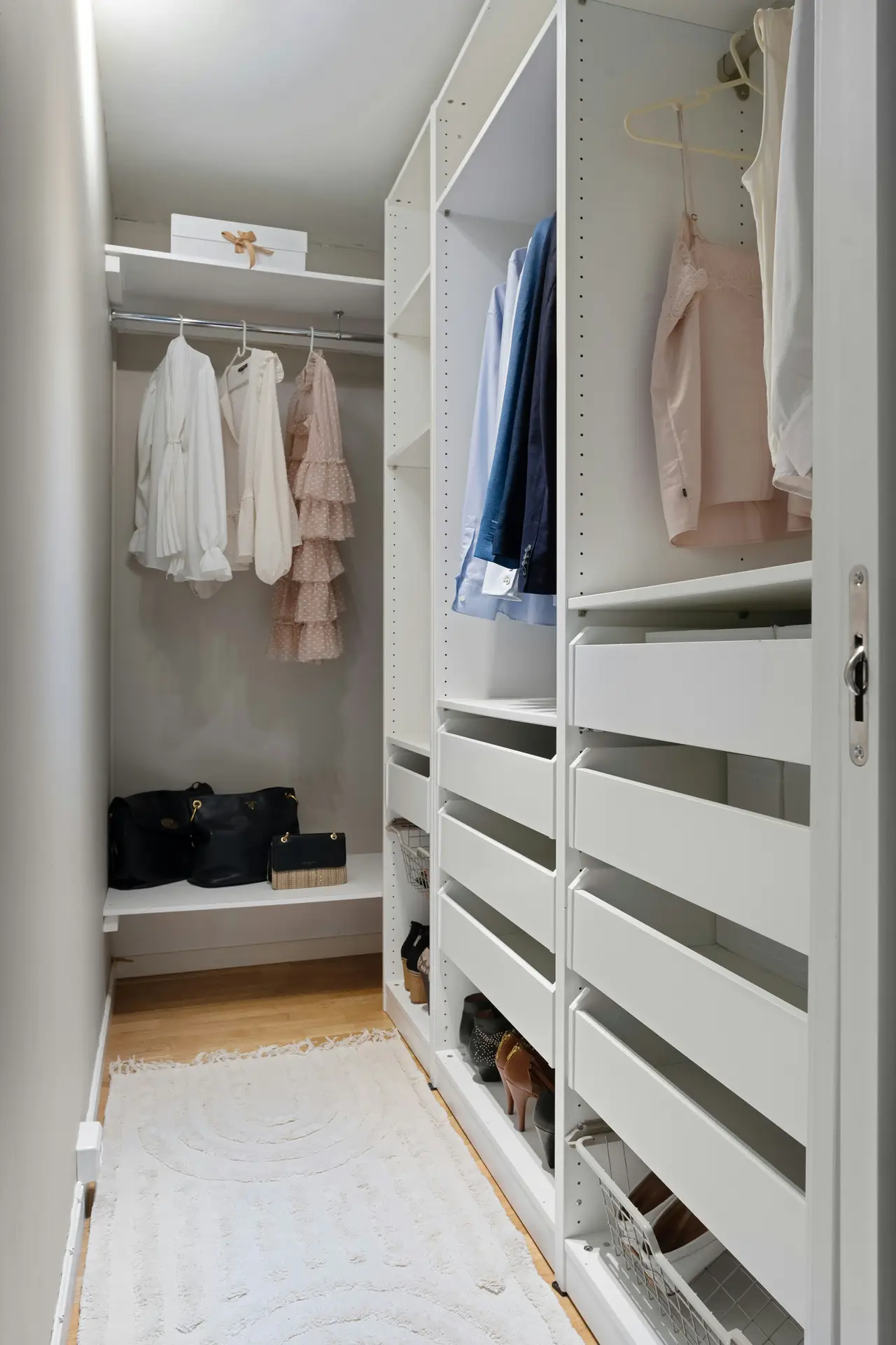 A bright minimal bedroom setup featuring a narrow built-in wardrobe with open shelving, hanging space, and organized drawers. Light clothing, handbags, and neatly arranged shoes create a clean, airy feel, while a soft white rug adds warmth to the streamlined storage area.